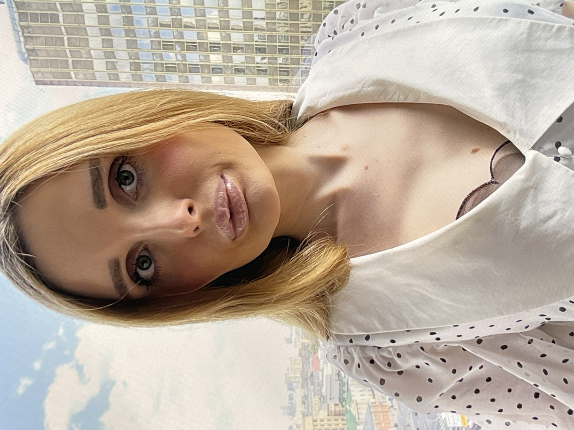 A young woman with blonde hair and fair skin is pictured in a close-up selfie. She is wearing a white blouse with black polka dots and a large collar. She has prominent makeup on her eyes and lips. Behind her, out of focus, is a cityscape with a tall building and a partly cloudy sky.