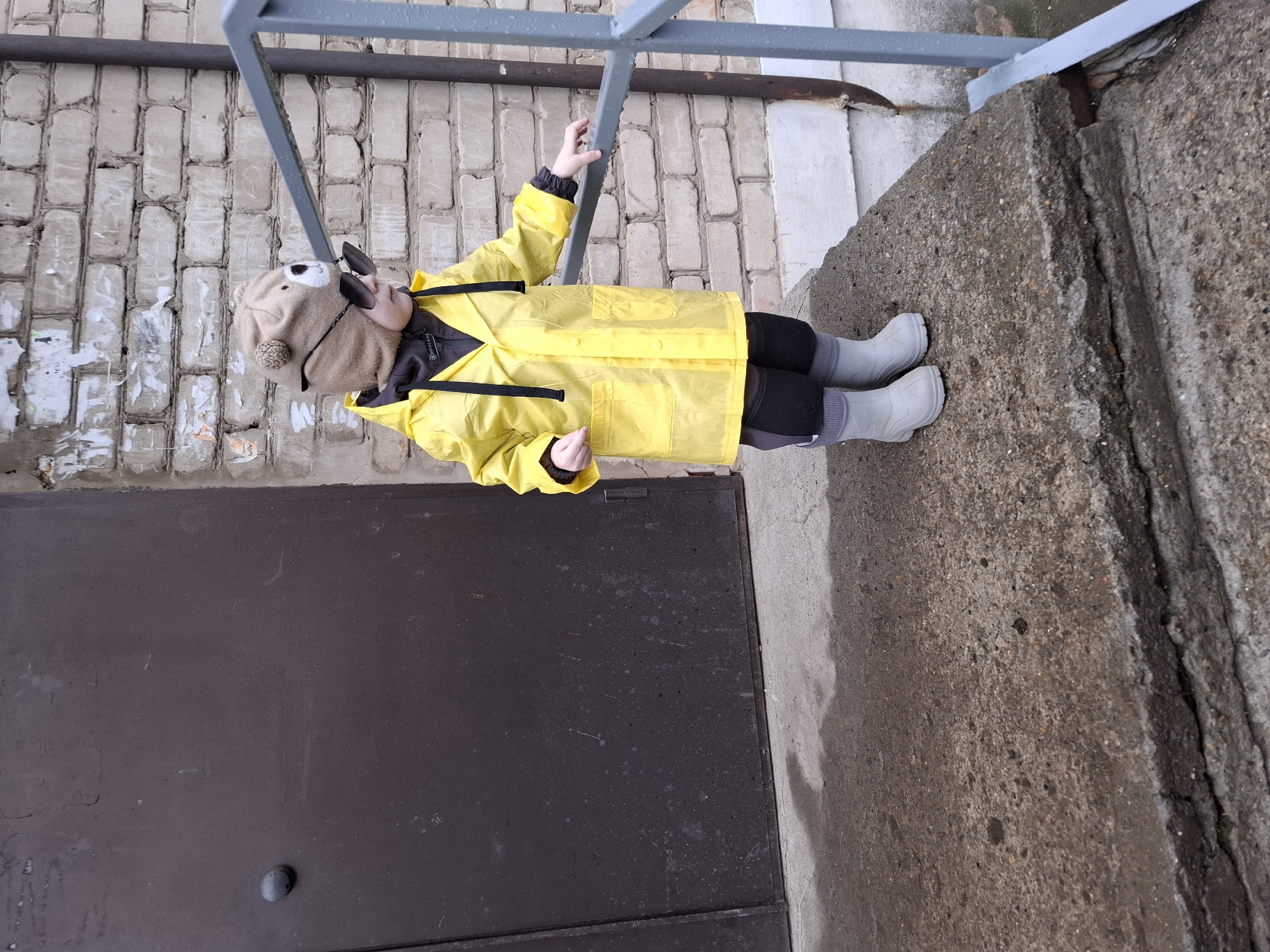 A young child, likely a toddler or preschooler, is depicted standing outdoors in Chaikovsky, Russia. The child is dressed in a bright yellow raincoat, dark leggings, and grey rubber boots with grey socks. They wear a light brown knitted hat resembling a bear, complete with ears and a face, and dark sunglasses.
The child is seen holding onto a grey metal railing with their right hand, looking slightly upwards. Their left hand is down by their side, with the palm facing up. The setting appears to be an urban or industrial area. Behind the child is an old, weathered brick wall with patches of white paint or plaster peeling off. To the child's left is a large, dark brown or black metal door or hatch, possibly for a utility access or storage, with a small dark knob visible towards the bottom. The ground beneath the child is rough, textured concrete, which shows signs of wear and possibly dampness.
The overall impression is of an overcast day, fitting for the child's protective attire of a raincoat and boots. There is no clear, legible text visible in the image apart from a small, unreadable tag on the child's inner clothing. The child's expression is not fully visible due to the sunglasses and angle, but their posture suggests engagement with their surroundings.