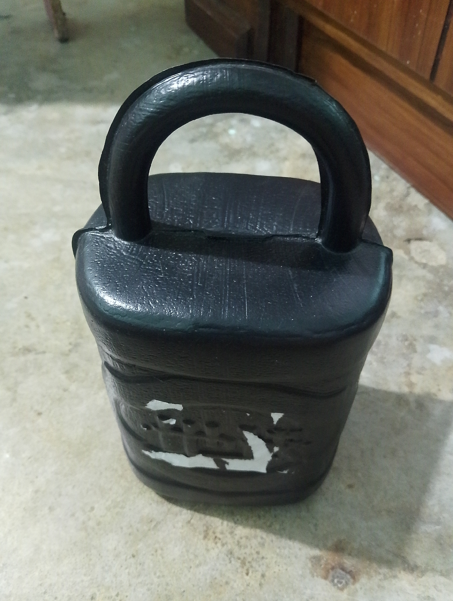 A black kettlebell sits on a concrete floor. The kettlebell is partially covered by a white sticker, and some of its texture is visible. Behind the kettlebell, a wooden cabinet or door is visible, and to the left, a portion of a chair leg can be seen. The overall environment suggests an indoor setting in Serbelawan, Indonesia.