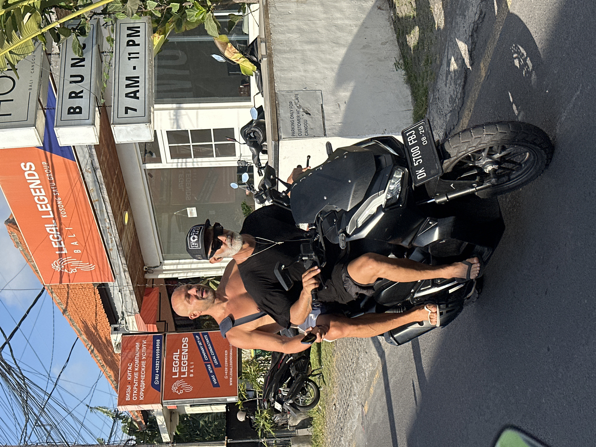 In North Kuta, Indonesia, two men are riding a black motorcycle on a paved road. The man in the back is wearing a black t-shirt, dark shorts, and a black baseball cap, with sunglasses and a necklace. The man in the front is shirtless, wearing white shorts and flip-flops, with his left arm around the driver. Both men are looking forward. The motorcycle has a license plate that reads "DK 5700 FBJ". In the background, a building with orange awnings and signs advertising "LEGAL LEGENDS BALI" and "BRUNCH 7 AM - 11 PM" is visible. There are several power lines overhead, and a motorcycle is parked on the side of the road. A sign on a wall in the background indicates "PARKING ONLY FOR CUSTOMER HO CAFE CANGGU".