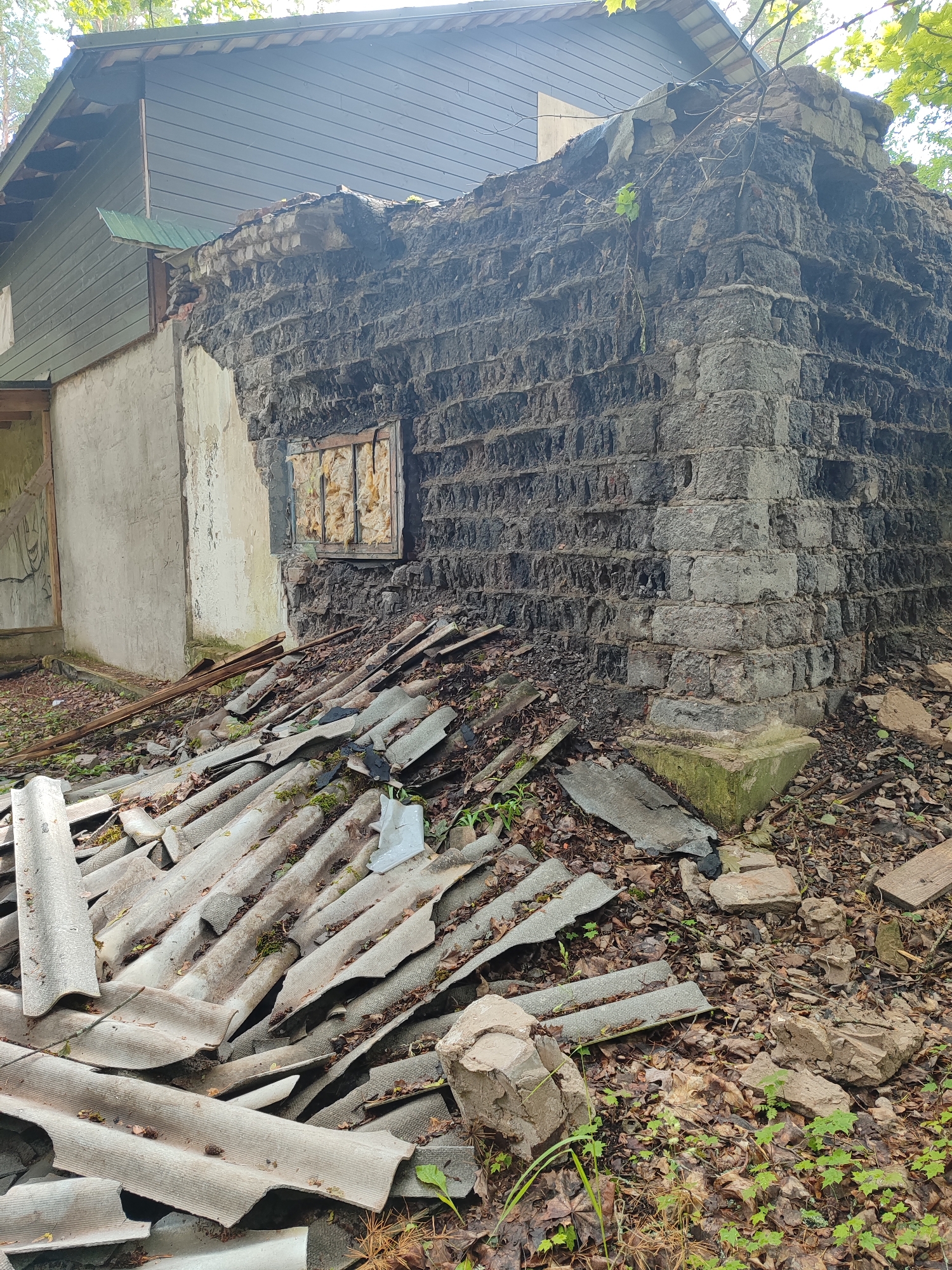 The image shows the exterior of a dilapidated building. A damaged brick wall, partially collapsed, dominates the right side of the frame, with exposed insulation visible in a recessed window opening. The wall appears to be made of large, irregular bricks. To the left of the brick wall, a lighter-colored, stuccoed wall is visible, with what appears to be graffiti on a wooden panel leaning against it. A pitched roof structure with dark siding and a section of green metal roofing extends from the top left of the image. The ground in front of the building is covered with debris, including broken pieces of corrugated roofing material, wood planks, leaves, and scattered stones. The environment appears to be outdoors, with trees and foliage visible in the background. The scene suggests neglect and disrepair.