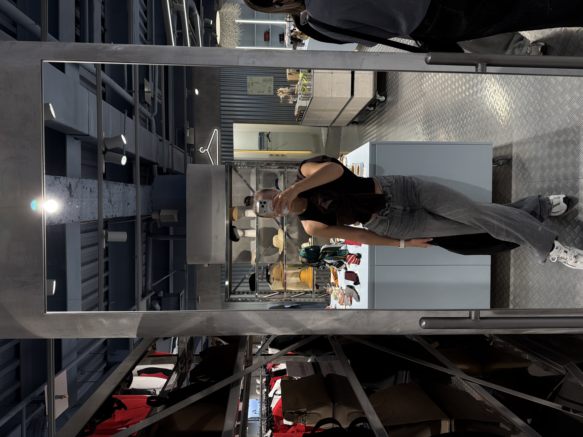 The image depicts a young woman taking a mirror selfie in what appears to be a retail store. She is wearing a dark top, grey jeans, and white sneakers. She is holding a smartphone in her right hand. In the reflection, the store's interior is visible, including shelves stocked with clothing and accessories, display counters, and industrial-style ceiling elements with exposed pipes and lighting fixtures. The floor is made of a diamond-plate metal material. The overall aesthetic suggests a modern, possibly trendy, boutique. The location is identified as a store in Moscow, Russia.