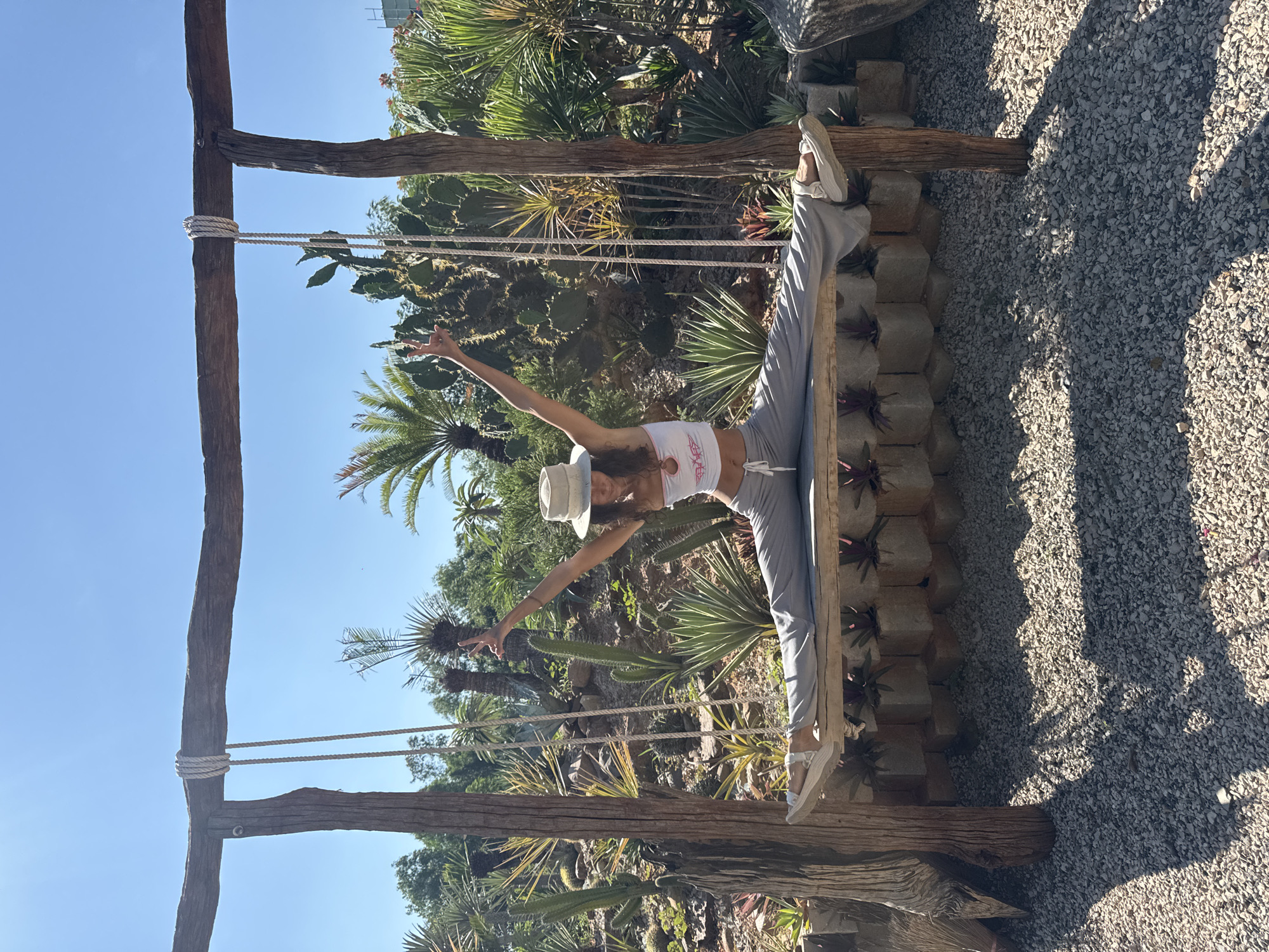 A young woman with long dark hair and a white hat is performing a split on a wooden swing in a garden in Mui Ne, Vietnam. She is wearing a white crop top and grey pants. The swing is suspended from a wooden frame by ropes. Behind the woman, the garden is filled with various plants, including cacti and palm trees, under a clear blue sky. The ground is covered in gravel.