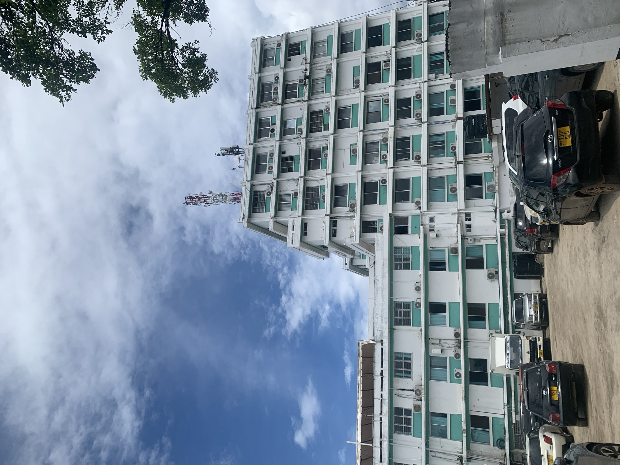 The image displays the exterior of Kairuki Hospital, located in Dar es-Salaam, Tanzania. The prominent subject is a multi-story building, primarily white with teal-colored panels bordering its numerous windows. The central section of the building rises to at least seven visible floors, exhibiting a stepped-back architectural design. Its rooftop features multiple communication antennas, including a distinct red and white lattice tower. Numerous external air conditioning units are mounted below most windows across the building's facade. In the foreground, an unpaved parking area is occupied by several vehicles. A white box truck, displaying "KAIRUKI HOSPITAL" in blue lettering, is positioned prominently. Other parked vehicles include black and silver passenger cars, some with visible Tanzanian license plates such as T924 DUJ and T710 DZD. A black water storage tank is situated on a stand near the base of the building, behind parked vehicles. The sky above is partly cloudy, showing a mix of blue sky and white cumulus clouds. Green foliage from trees is visible in the upper right quadrant of the image. No individuals are present in the scene.