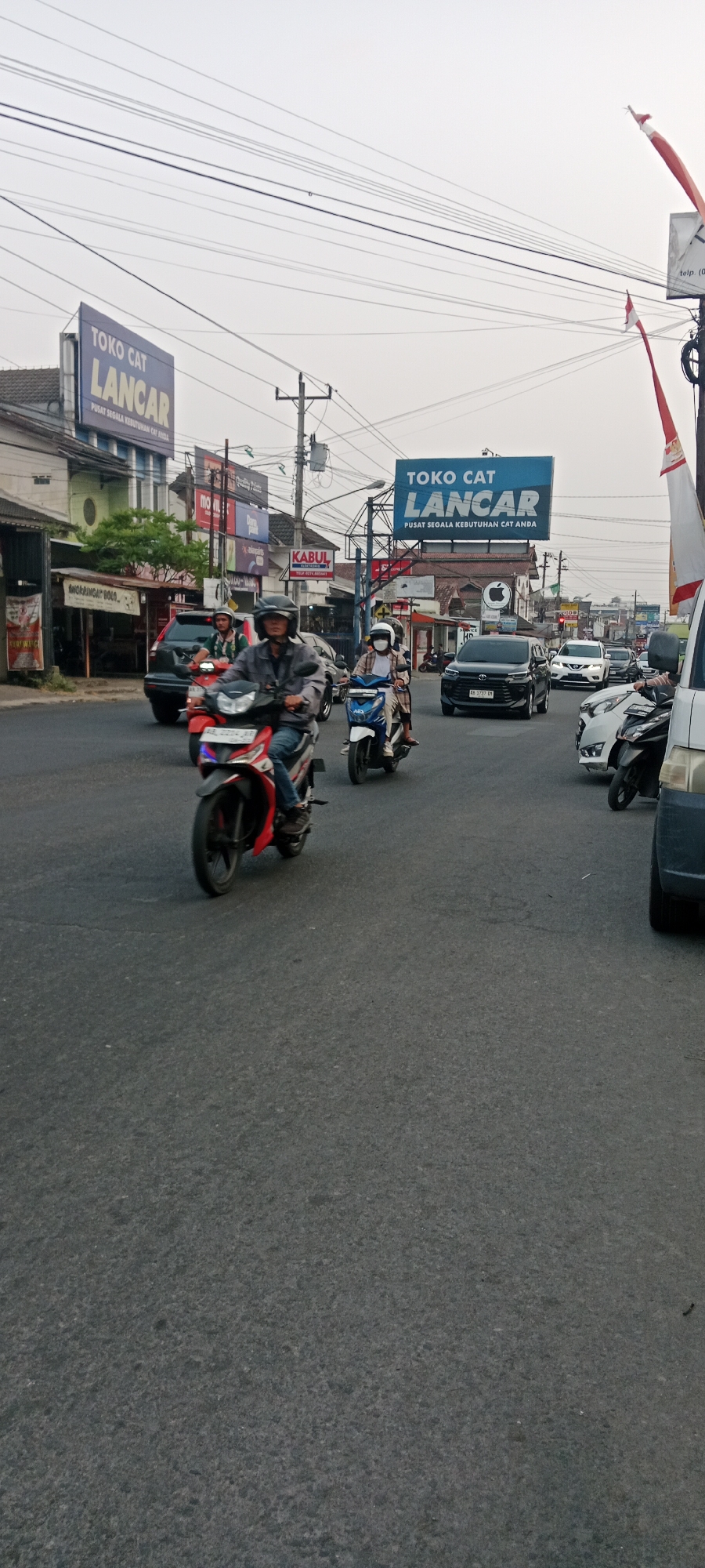 This is a street-level, full-shot photograph taken in Maguwoharjo, Indonesia. The image captures a busy road with several vehicles and pedestrians. In the foreground, a person is riding a red motorcycle. Behind them, another person is on a white and blue motorcycle, and further back, a person on a scooter is visible. Multiple cars, including a black SUV and a white sedan, are also present on the road. Buildings line the street, with prominent signage indicating businesses such as "TOKO CAT LANCAR" (Paint Store Lancar) and "KABUL ELEKTRONIK" (Kabul Electronics). Several utility poles and overhead wires crisscross the sky, which is overcast. Two Indonesian flags, red and white, are visible, one on a pole to the right and another hanging. The overall scene depicts a typical urban traffic environment in Indonesia, with a focus on the movement of vehicles and the commercial activity along the roadside.