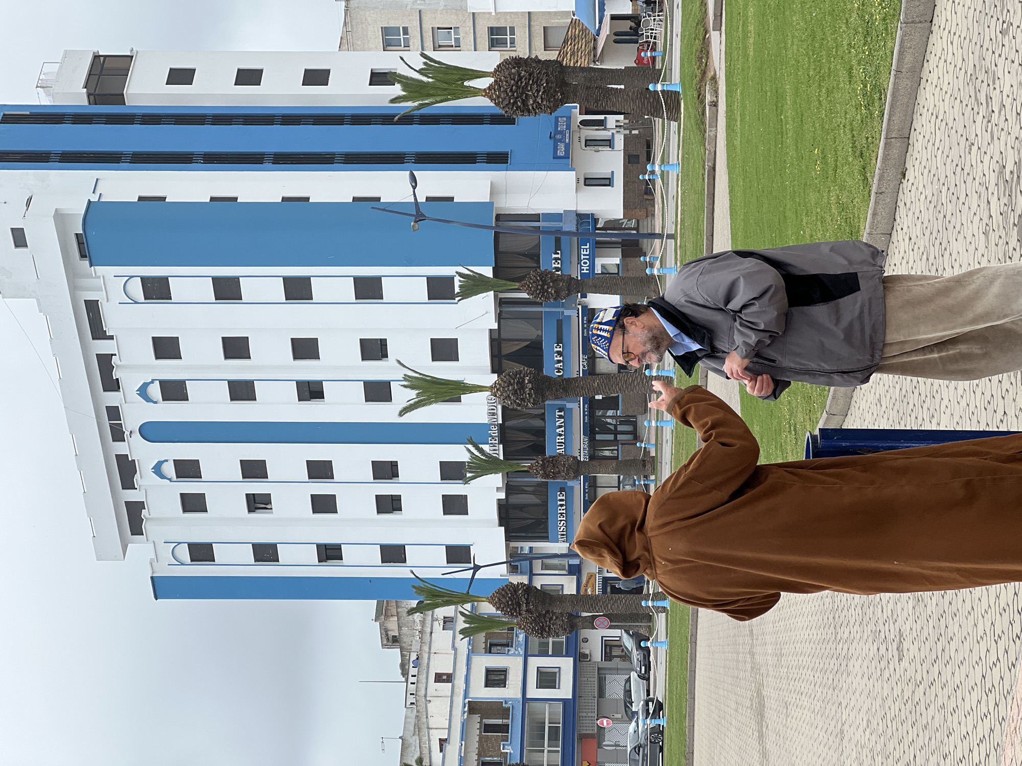 Two men interact on a paved walkway in M'Diq, Morocco, under an overcast sky. In the foreground, a man wearing a brown hooded robe (possibly a djellaba) stands with his back to the viewer, his right arm raised and hand gesturing towards the other man. The second man, facing partially away from the camera, wears a grey jacket, light brown or beige corduroy trousers, and a distinctive blue and white patterned cap. He looks downwards, seemingly at something in his hands or at the ground.
Behind the men, a prominent multi-story building dominates the midground. It features a modern design with white walls and horizontal blue bands running across its facade. Numerous dark, square windows punctuate the white sections. Several palm trees with thick, rough trunks are planted along the front of this building, partially obscuring the lower floors.
Visible text on the building indicates its functions: "HOTEL," "CAFE," "RESTAURANT," and "PATISSERIE." A partially visible sign below the "RESTAURANT" section appears to read "PLAGE DE MDIO," suggesting a beachside location. To the right of the paved area where the men stand, there is a neatly maintained grassy strip bordered by a curb. In the background, other residential or commercial buildings can be seen, some also featuring white and blue elements, and several parked cars are visible on the street. The overall scene suggests a typical daytime street or promenade setting in a coastal town.