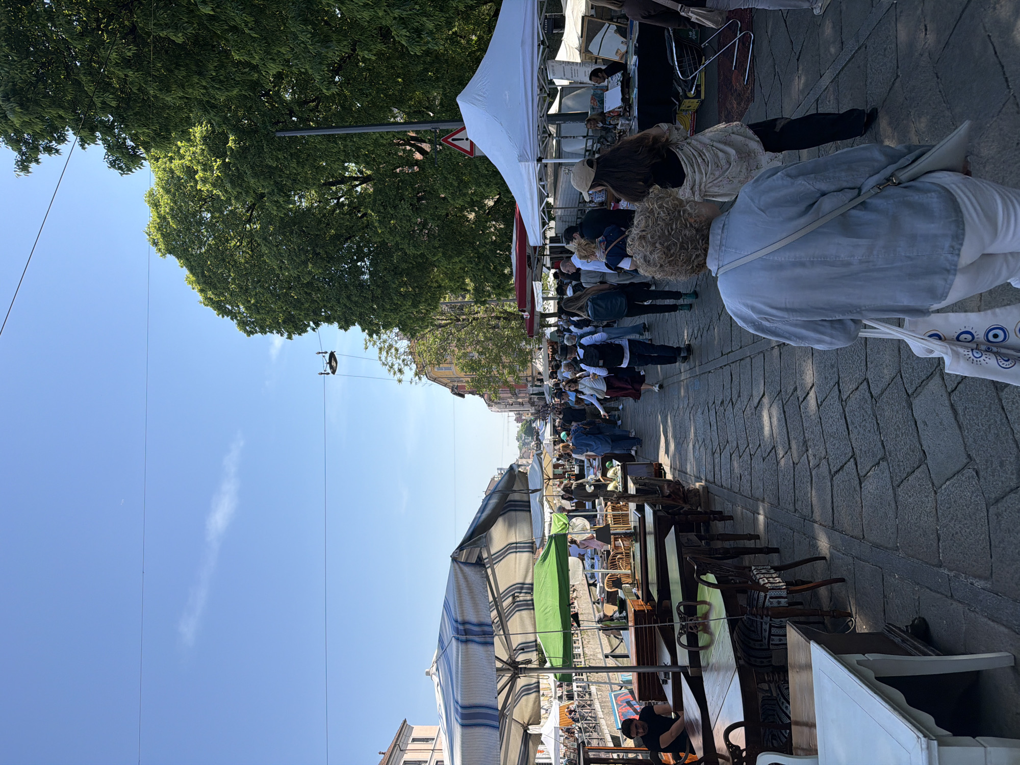 The image depicts a street scene in Milan, Italy, likely at a market. A crowd of people is walking along the cobblestone pavement between rows of vendor stalls. A large tree with dense foliage is visible on the left side of the image. Above the scene, a clear blue sky is visible, and power lines cross the sky. Along the right side of the image, there are chairs and tables. There are awnings.