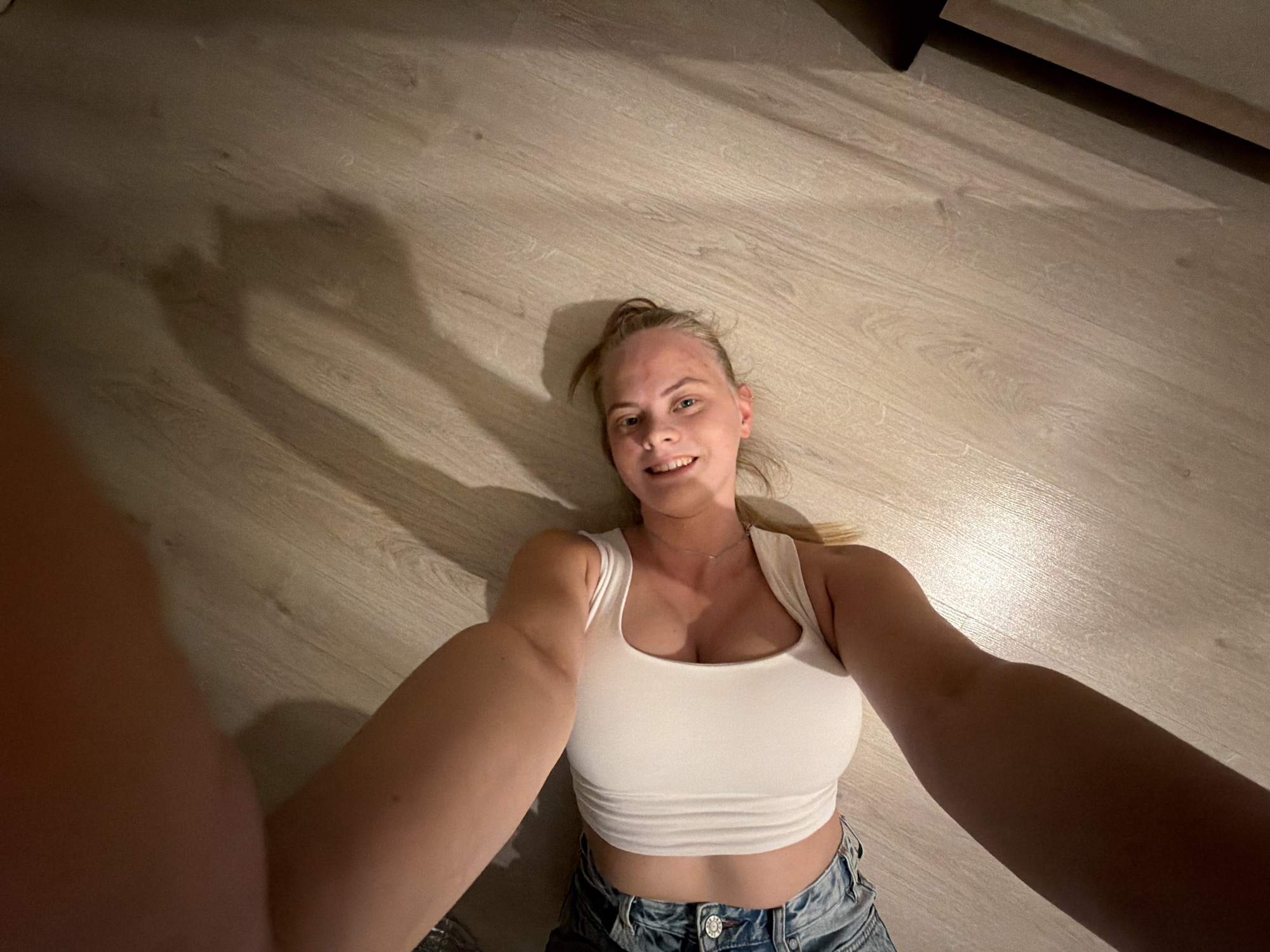 A young woman with blonde hair pulled back is lying on a light-colored wood-look floor, smiling at the camera. She is wearing a white tank top and denim shorts. Her arms are extended towards the camera, creating a selfie perspective. The lighting is soft, casting shadows on the floor. A dark object is visible in the upper right corner of the image. The location is impossible to determine based on this image.