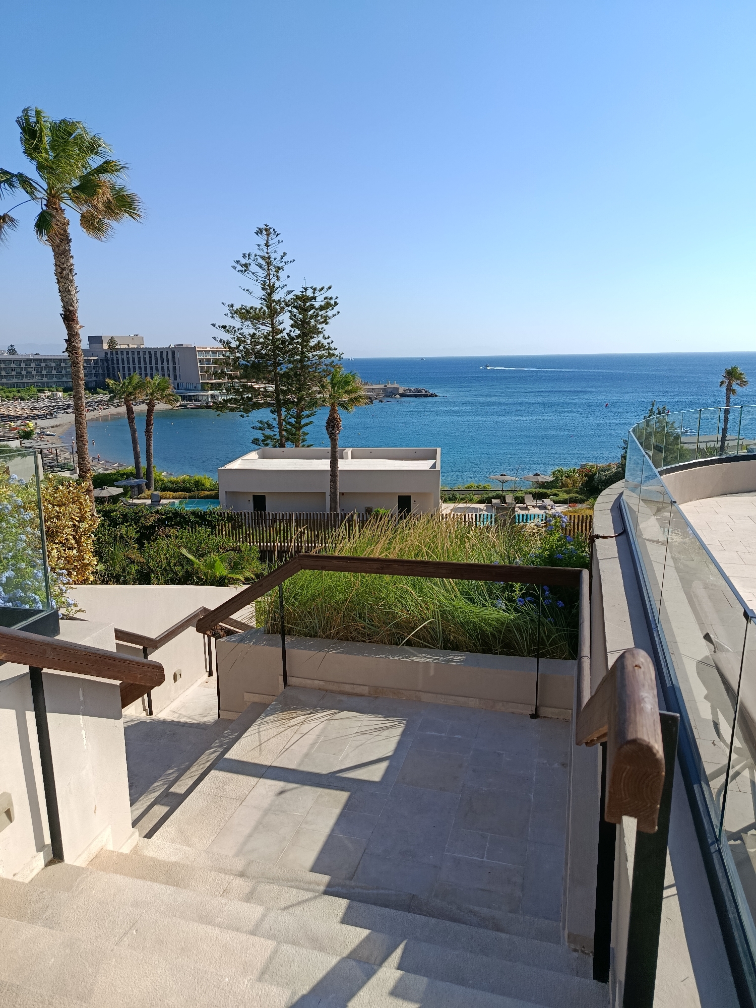 This is a wide, vertical, eye-level, outdoor shot taken from a balcony, looking down a set of concrete stairs towards a bay on a sunny day. The stairs lead down and to the left, with a railing made of dark wood and metal on the left side and a glass railing on the right side, separated by a planter filled with tall green grass. In the middle ground, a low-rise building with a white facade and dark windows is visible. Beyond that, palm trees and lush green vegetation are abundant. A swimming pool with lounge chairs is also visible. In the background, a wide expanse of blue water stretches to the horizon, with a sandy beach and a large hotel complex in the distance. The sky is clear and blue, with a faint hint of a boat on the water. The scene is likely a resort or hotel in the Municipal Unit of Rhodes, Greece, possibly in the city of Rhodes itself, given the presence of a large hotel and a bay. There are no people visible in the frame.