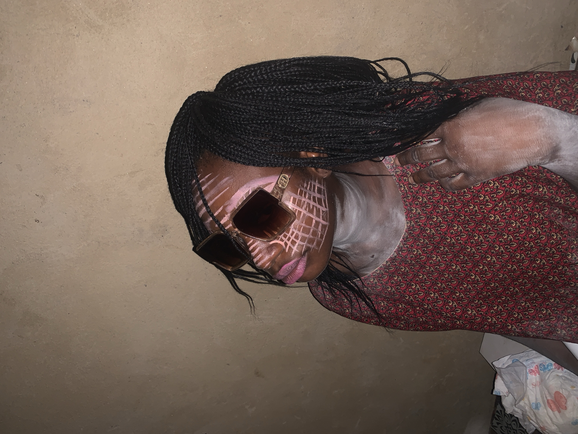 The image shows a person lying down, possibly on the floor, with their head turned to the side. The person has dark braided hair and is wearing sunglasses with a gold frame. Their face has white markings that resemble a grid pattern, and their lips are painted a bright pink. The person is also wearing a red dress with a floral pattern. Their right hand is visible and appears to be dusted with a white powder. The background is a plain, textured surface, likely a wall or floor, in a neutral color. A small portion of what appears to be a diaper or baby wipes is visible in the bottom right corner. The lighting suggests it is indoors or during a time of day with limited natural light. The overall impression is of a stylized portrait or artistic expression.