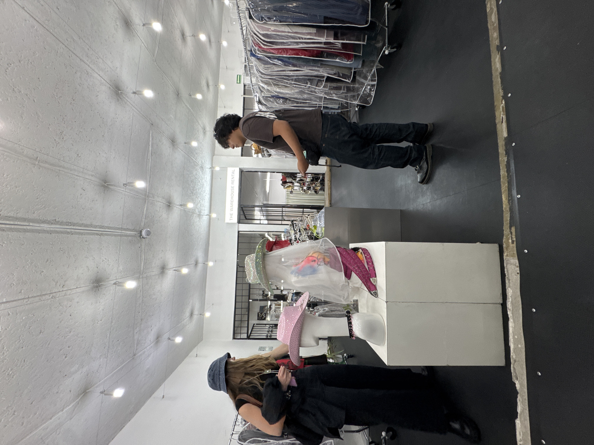 The image depicts the interior of a retail space in Mexico City, Mexico. The location appears to be a costume rental or sales business, given the prevalence of costumes and related items.
A man is standing near a counter examining something, possibly merchandise or a display. He is wearing a dark shirt and jeans. Another person, possibly female, is kneeling near mannequins dressed in costumes. She has long hair and is wearing a denim hat and a dark jacket.
There are several mannequins displaying various types of hats and costumes. Clothing racks filled with garments in protective plastic covers are visible in the background. The interior has bright, even lighting with overhead lamps. The floor is dark, and the walls are light-colored. A sign reading "THE WAREHOUSE RENTAL" is noticeable in the background.