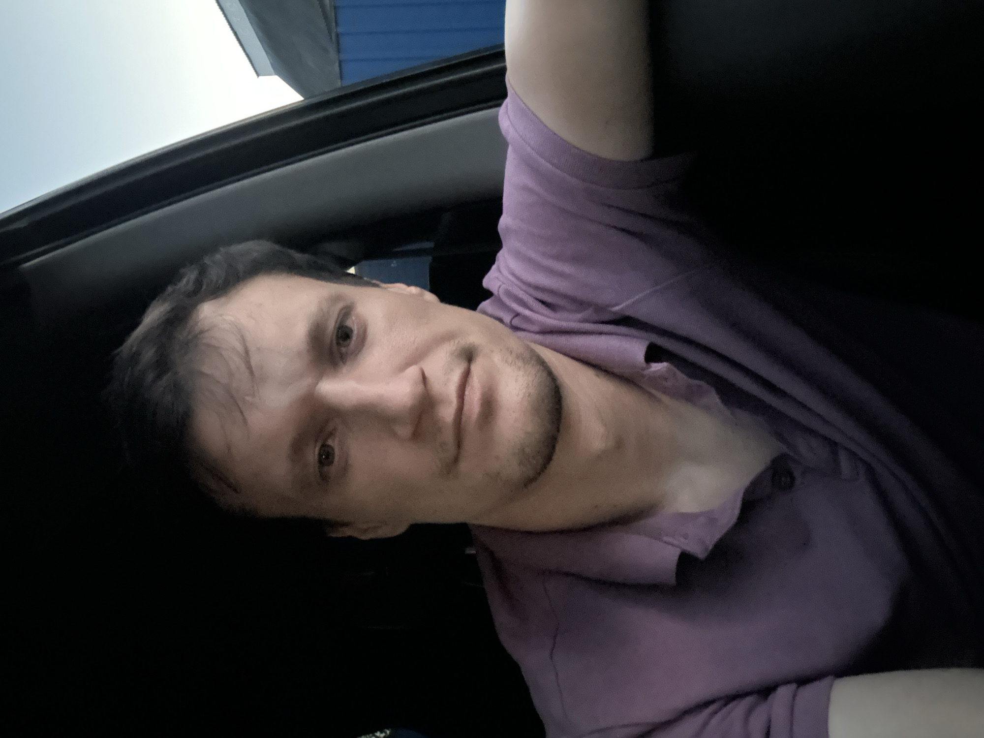 A medium shot, taken from inside a vehicle, shows a man with dark hair and brown eyes. He is wearing a purple polo shirt. The man is looking directly at the camera. The background shows a blurred blue building and a pale sky, suggesting an outdoor setting, possibly in Shatava, Ukraine. The lighting is dim, with shadows present.