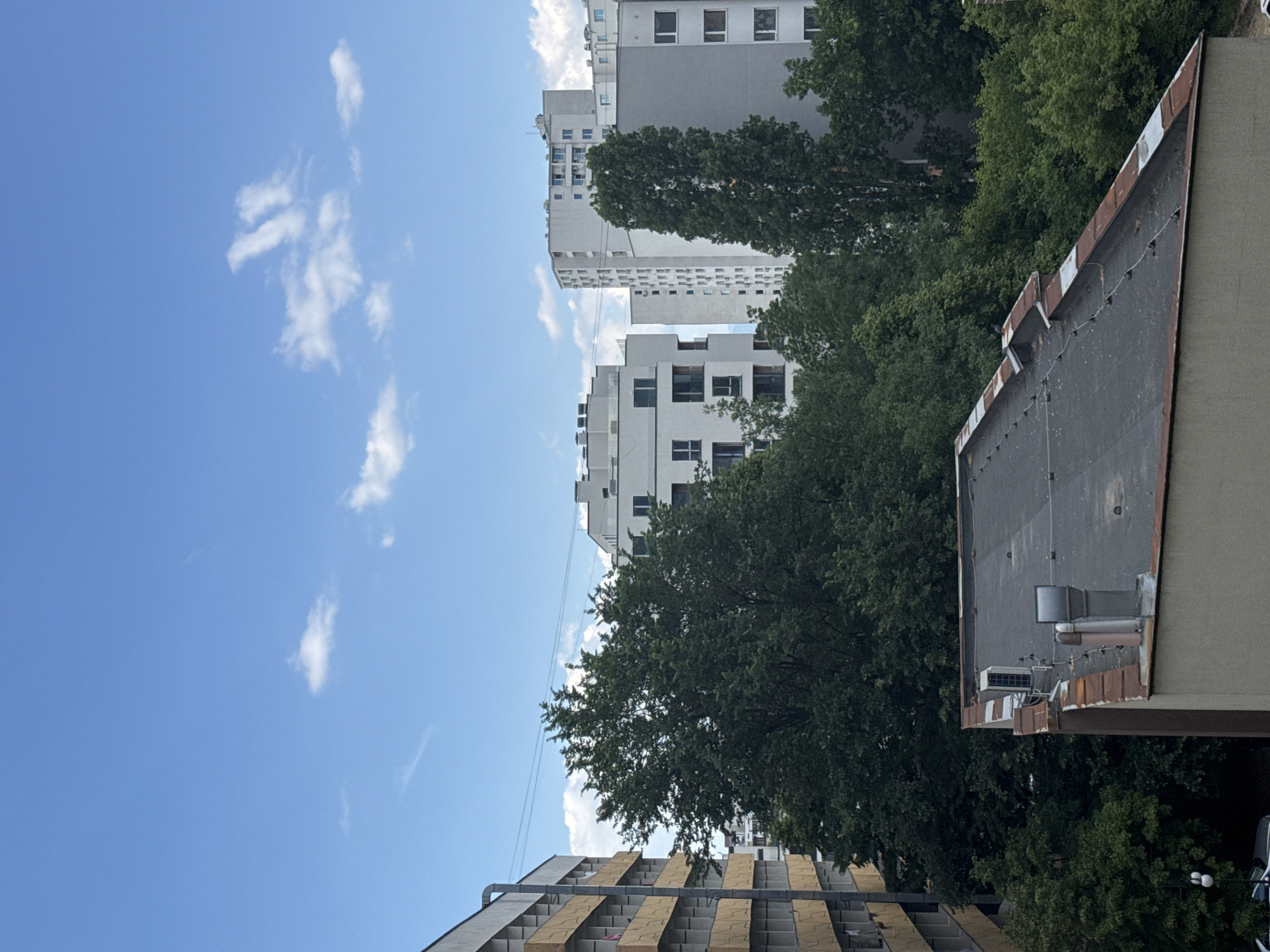 The image shows a view in Sofia, Bulgaria, looking up at buildings and a blue sky with some white clouds. Several multi-story apartment buildings are visible, interspersed with green trees. The roofs of adjacent buildings are also partially visible, one showing a flat roof with some vents and mechanical equipment, the other showing a terrace style with beige tiles. The sky is bright blue with scattered, fluffy white clouds.