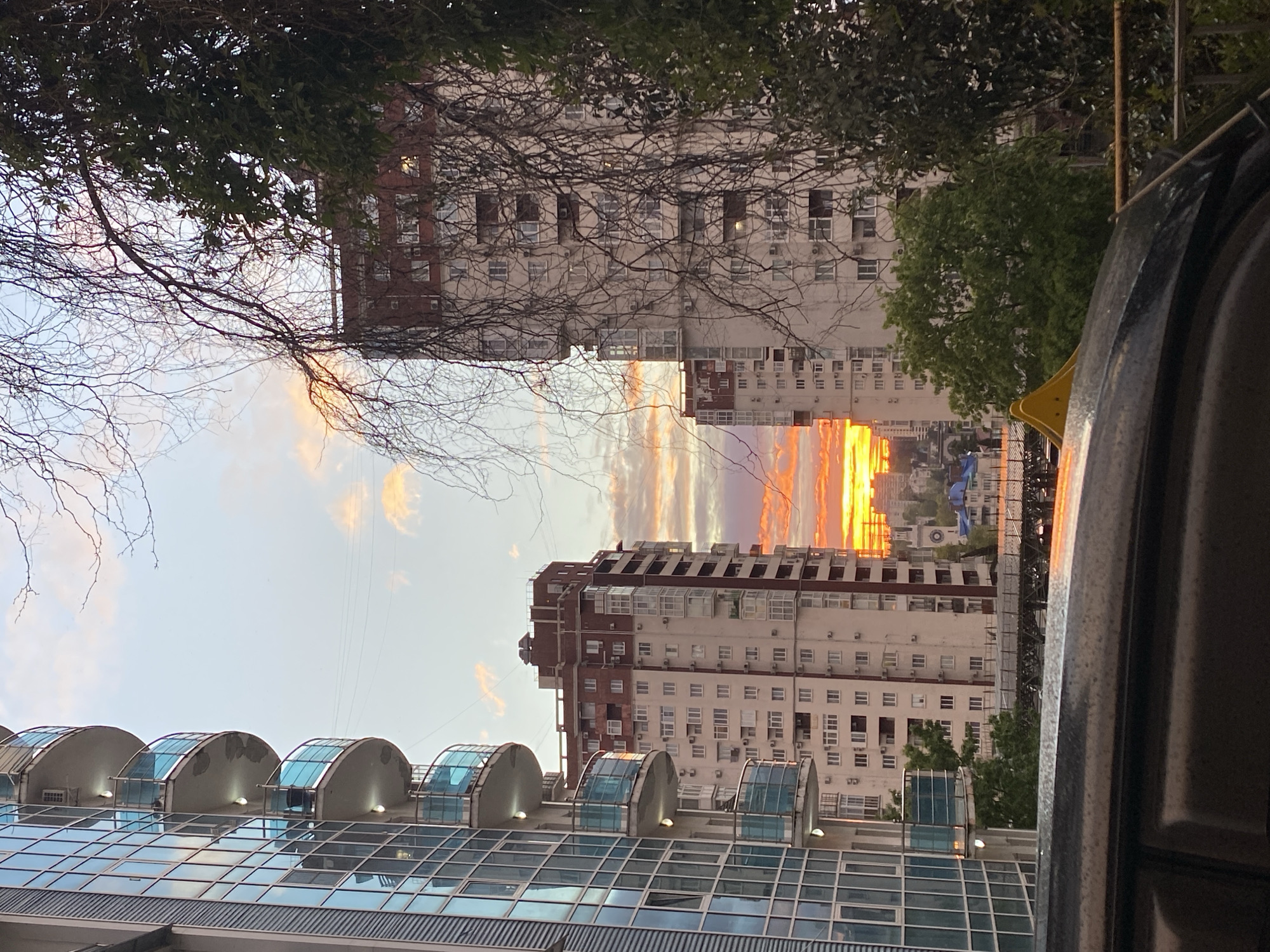 The image captures a sunset scene between tall buildings. The buildings are primarily white with some brown brick accents, featuring numerous windows and what appear to be air conditioning units. A building in the foreground has a modern architectural style with a glass and metal exterior featuring semi-circular bay windows. Trees and bare branches are visible, framing the top of the image. In the center, the sky glows with orange and purple hues of the setting sun. The setting appears to be urban. No people are visible. There is no way to determine the location is in the city of Sochi, Russia from the image alone.