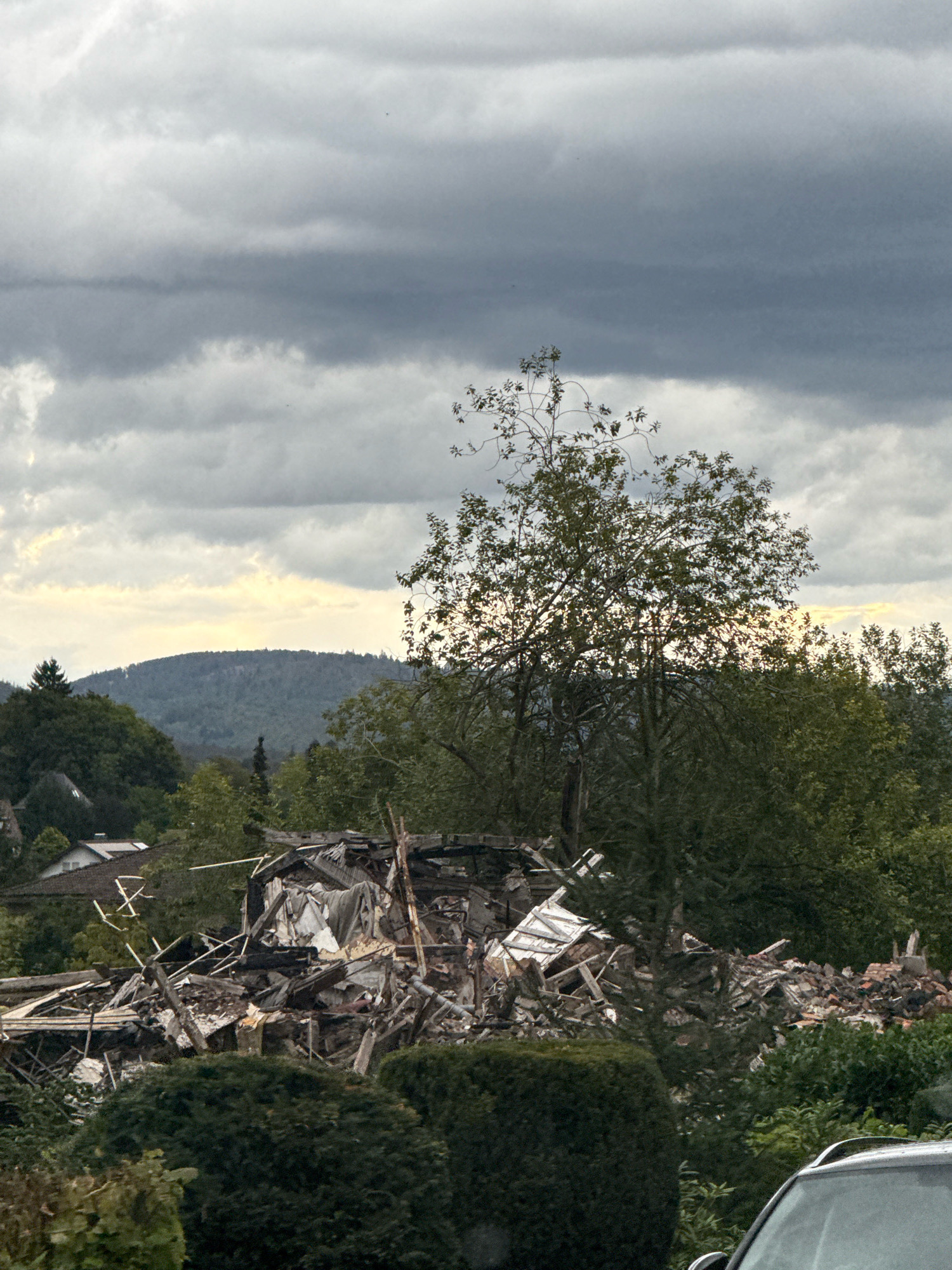 The image depicts the aftermath of a building collapse in Bad Sachsa, Germany. The scene is dominated by a large pile of rubble, consisting of broken wood, metal, bricks, and insulation materials, suggesting structural failure. Debris is scattered across the ground, with some larger structural elements, like beams and pieces of roofing, still identifiable. A large tree with sparse leaves stands prominently in front of the rubble pile, its branches partially obscuring the view. In the background, a gentle hill covered in trees rises under a heavily overcast sky with streaks of pale yellow light visible near the horizon. Several houses with tiled roofs are visible in the mid-ground, nestled among dense green foliage. In the foreground, the top of a dark-colored car is visible on the right side, indicating its presence at the scene. The environment appears to be a semi-rural area, with residential buildings interspersed with significant vegetation. There are no visible people or animals in the image.