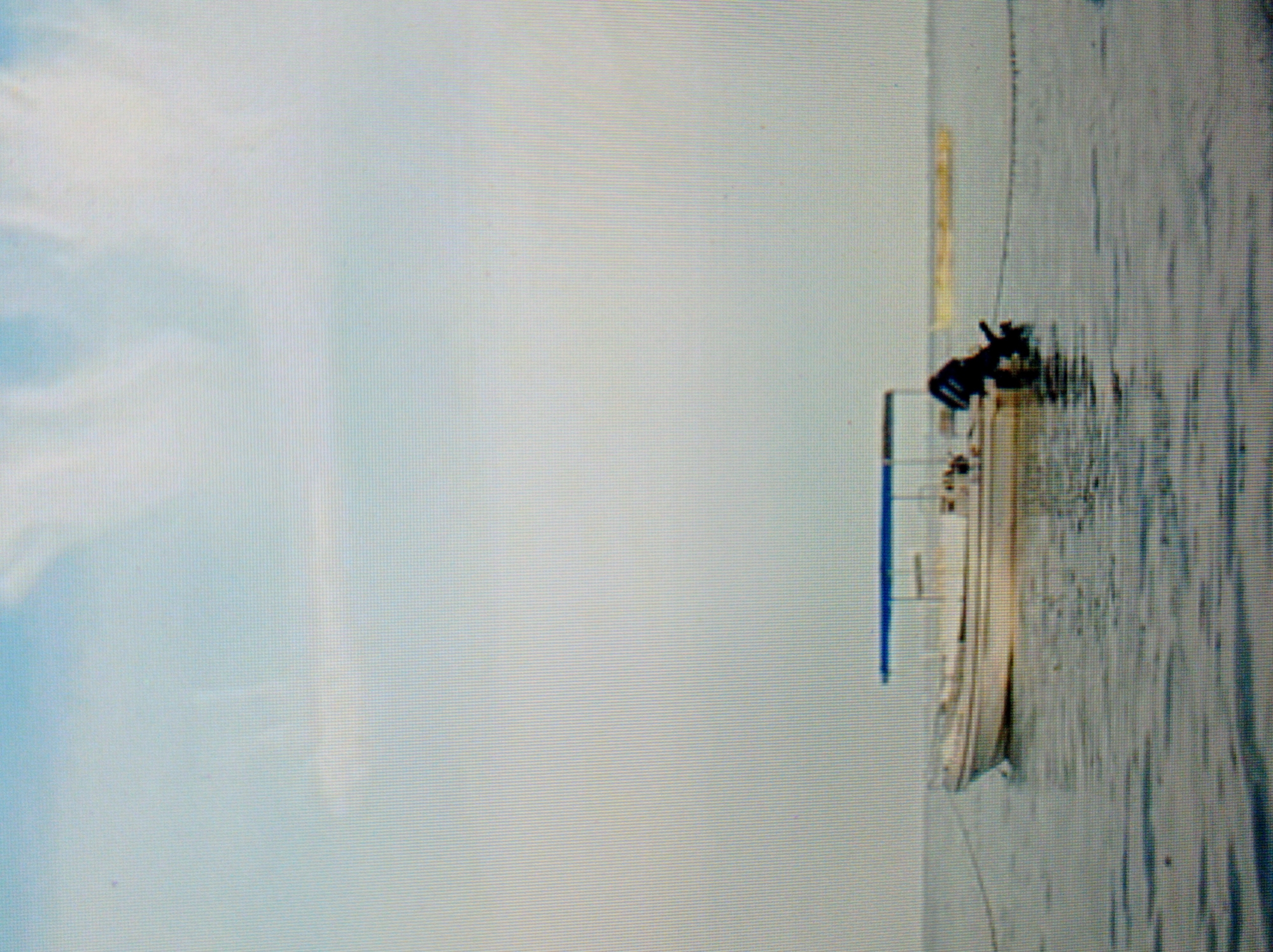A wide shot shows a white pontoon boat with a blue canopy floating on calm, gray water under a hazy sky. The boat is anchored by a rope to something off-camera to the left and also appears to be tethered to a floating yellow object in the distance to the right. The boat has an outboard motor at the stern. The image is low-resolution and appears to be a photograph taken of a screen. There is no discernible indication of the city of Mukachevo, Ukraine, in this image.