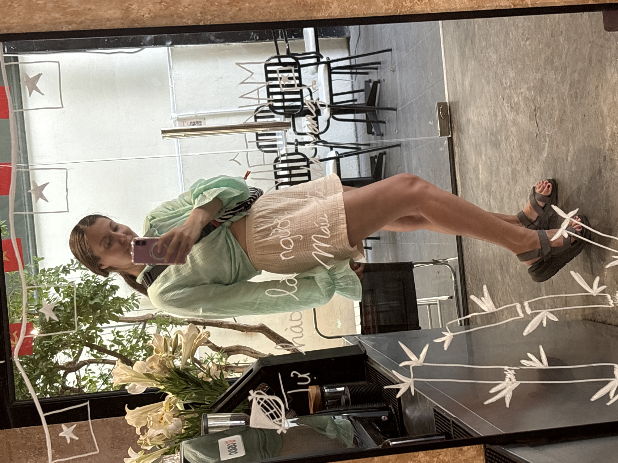 A mirror selfie shows a woman in a light green top and tan shorts, holding a purple smartphone. The reflection includes a storefront with chairs and tables, foliage, white flowers, and a counter with coffee equipment. The reflection is overlaid with decorative drawings, including Vietnamese flags and bamboo. The woman is wearing gray sandals. The location appears to be in Nha Trang Ward, Vietnam, based on text visible in the reflection.