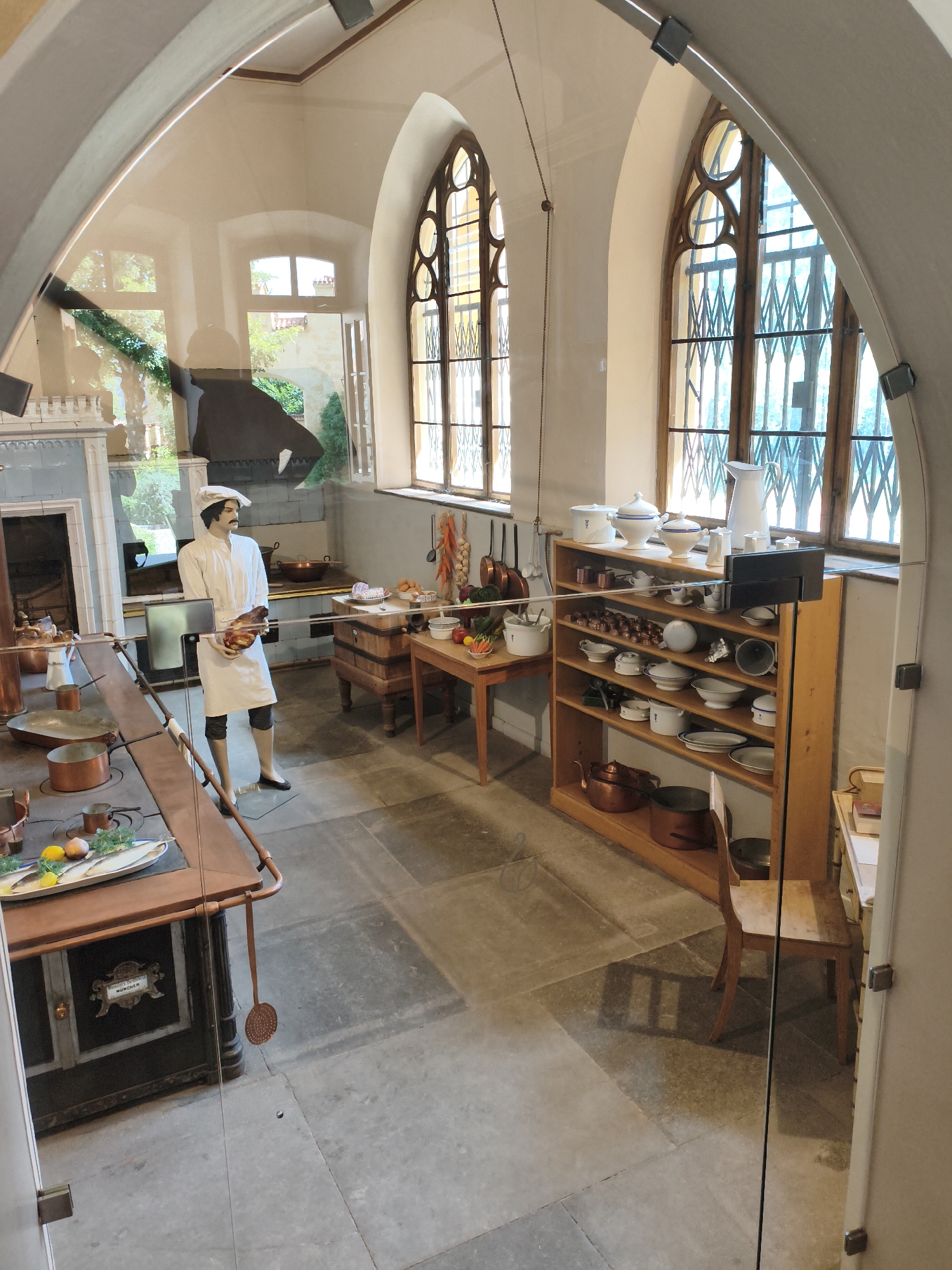 This is a forensic, matter-of-fact description of a mannequin in a historical kitchen exhibit, likely located in the Hohenschwangau Castle in Schwangau, Germany.
The scene depicts a spacious, well-lit kitchen interior. In the center, a male mannequin dressed as a chef stands holding a large piece of meat. The mannequin is wearing a white chef's uniform, including a hat, coat, and apron, with dark stockings and black shoes. The kitchen is equipped with a large, dark stove with copper pots and pans on its surface and nearby. To the right of the mannequin is a wooden preparation table laden with various kitchen utensils, vegetables, and other food items. Further to the right, a tall, wooden shelving unit is filled with a collection of white ceramic dishes, bowls, pitchers, and smaller kitchen tools. A wooden chair is positioned in front of the shelving unit. The room features large, arched windows with intricate wooden lattices, allowing natural light to enter. The floor is made of large, gray stone slabs. The overall impression is of a meticulously preserved historical kitchen environment.