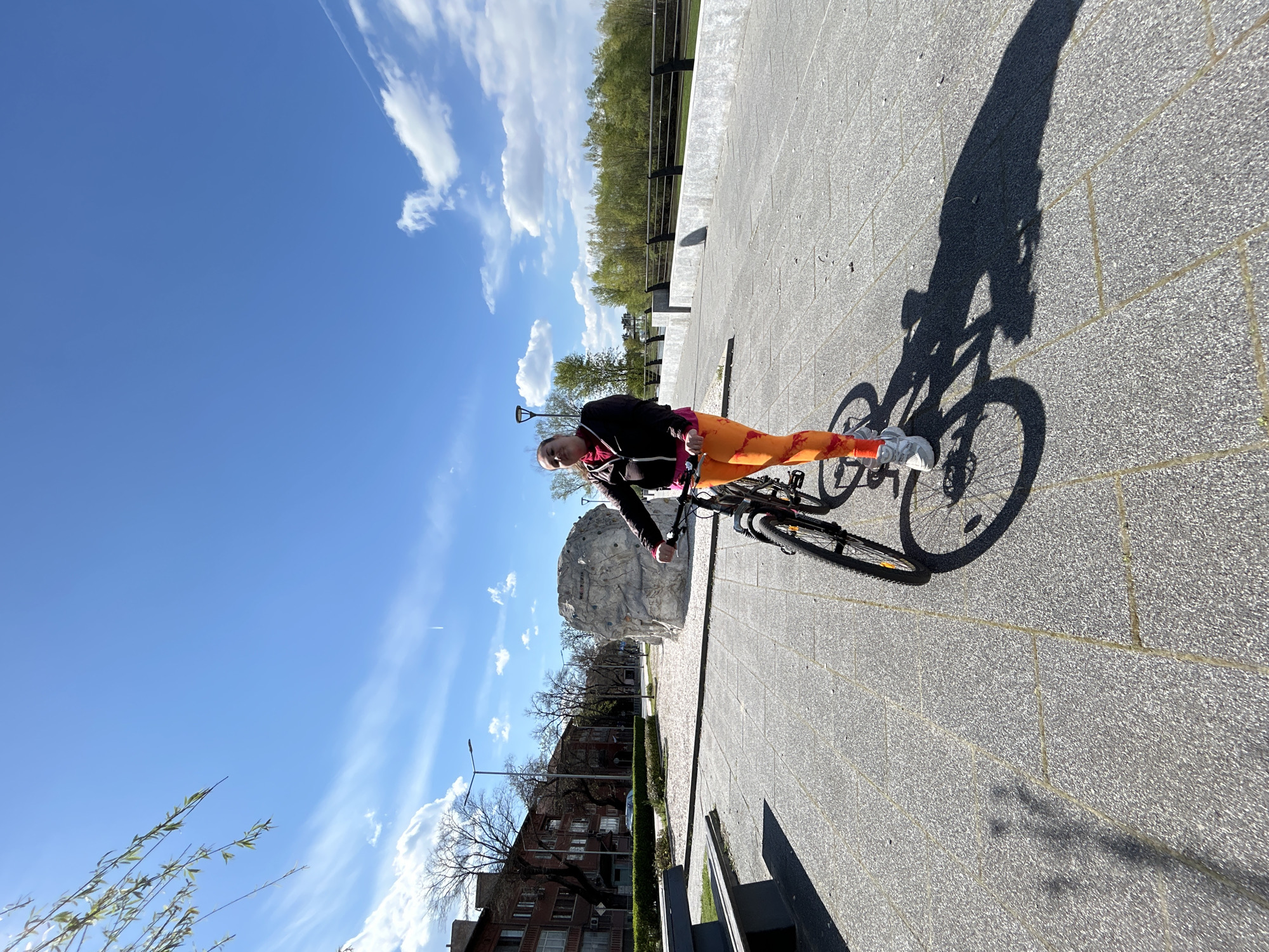 A person stands with a bicycle on a paved surface on a sunny day. The person is wearing orange leggings, a black jacket, and white sneakers. The shadow of the person and the bicycle is cast on the pavement. In the background are trees, buildings, and a blue sky with clouds. The location is Plovdiv, Bulgaria, near the stone sculpture, likely along the Maritsa River.