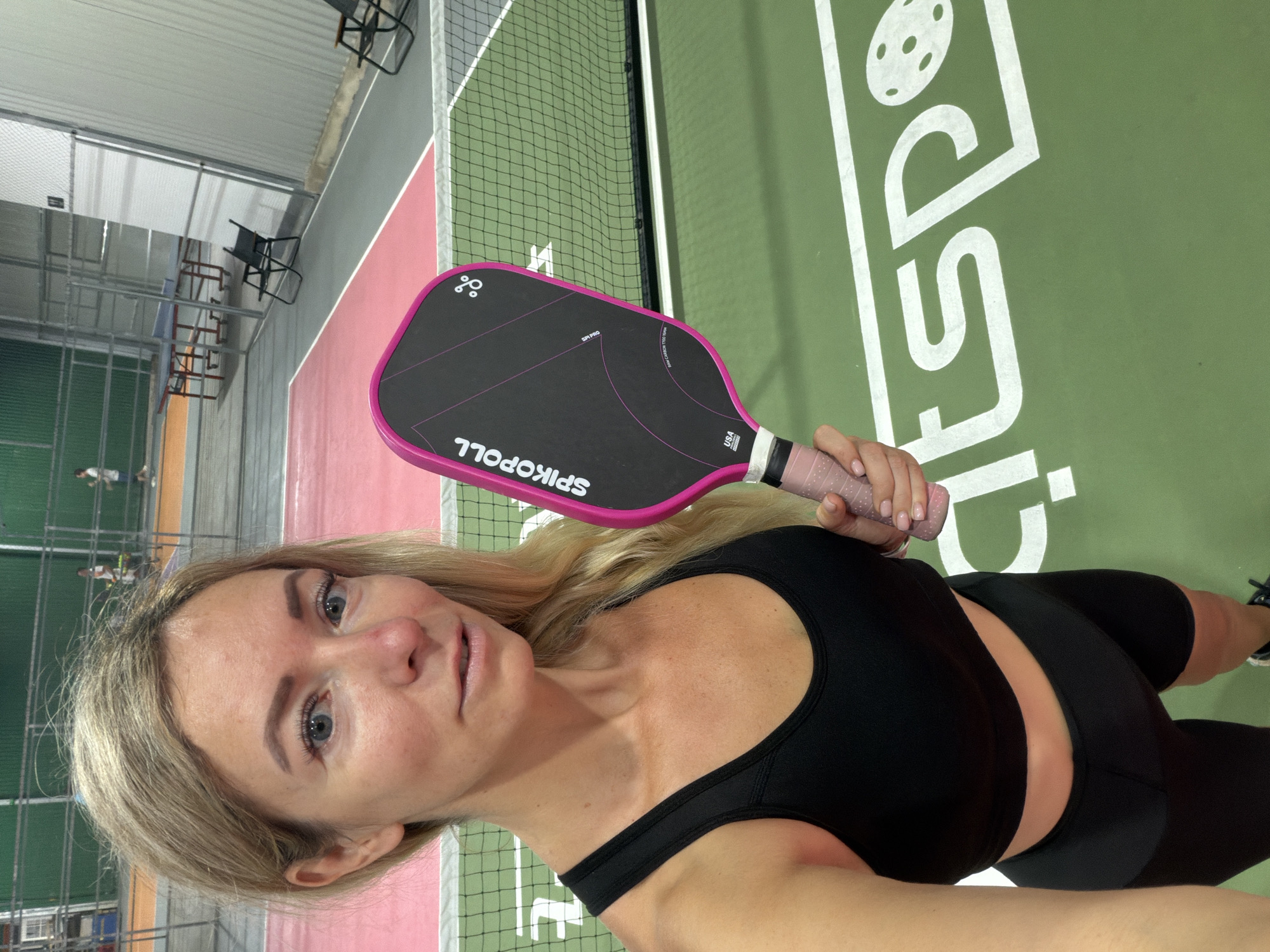 A woman is taking a selfie in an indoor sports facility. She is wearing a black sports bra and black shorts, and she is holding a pink pickleball paddle. The facility has green and pink courts, and there are nets and chairs visible in the background. The woman has blonde hair and is smiling at the camera. There is no indication that this location is in Da Lat, Vietnam.
