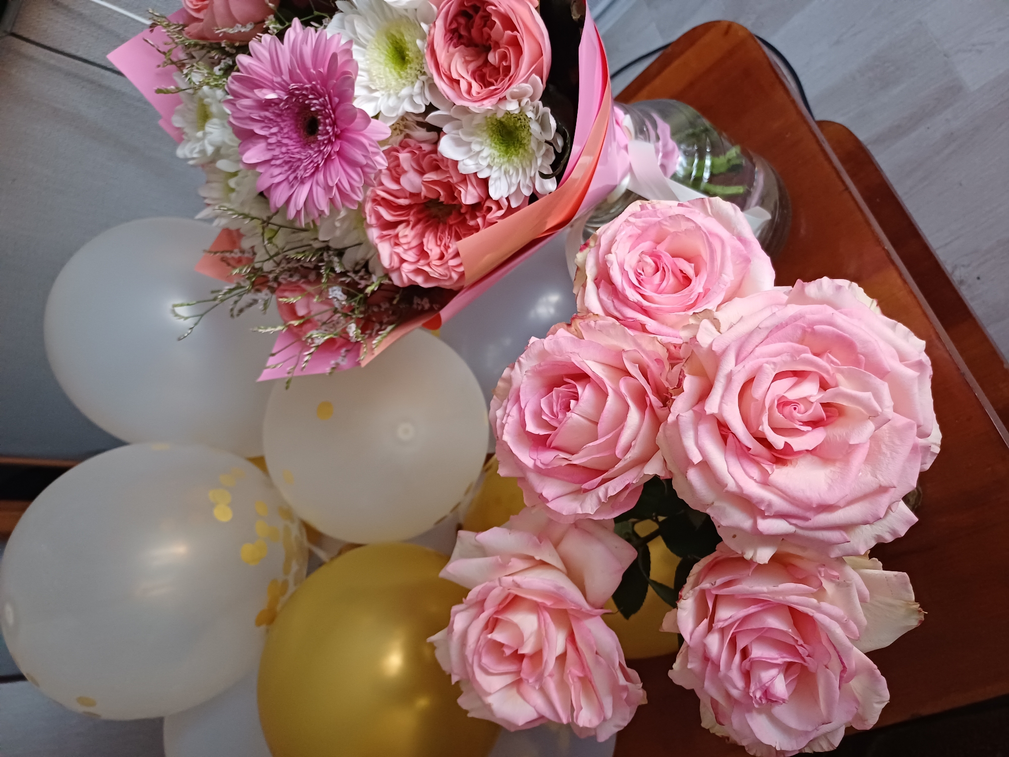 A bouquet of pink roses and other flowers is displayed on a wooden surface. The bouquet is in a glass vase tied with a pink ribbon. In the background, there are several balloons, including white ones with gold confetti and a gold one. The setting appears to be indoors, possibly a home or event space.