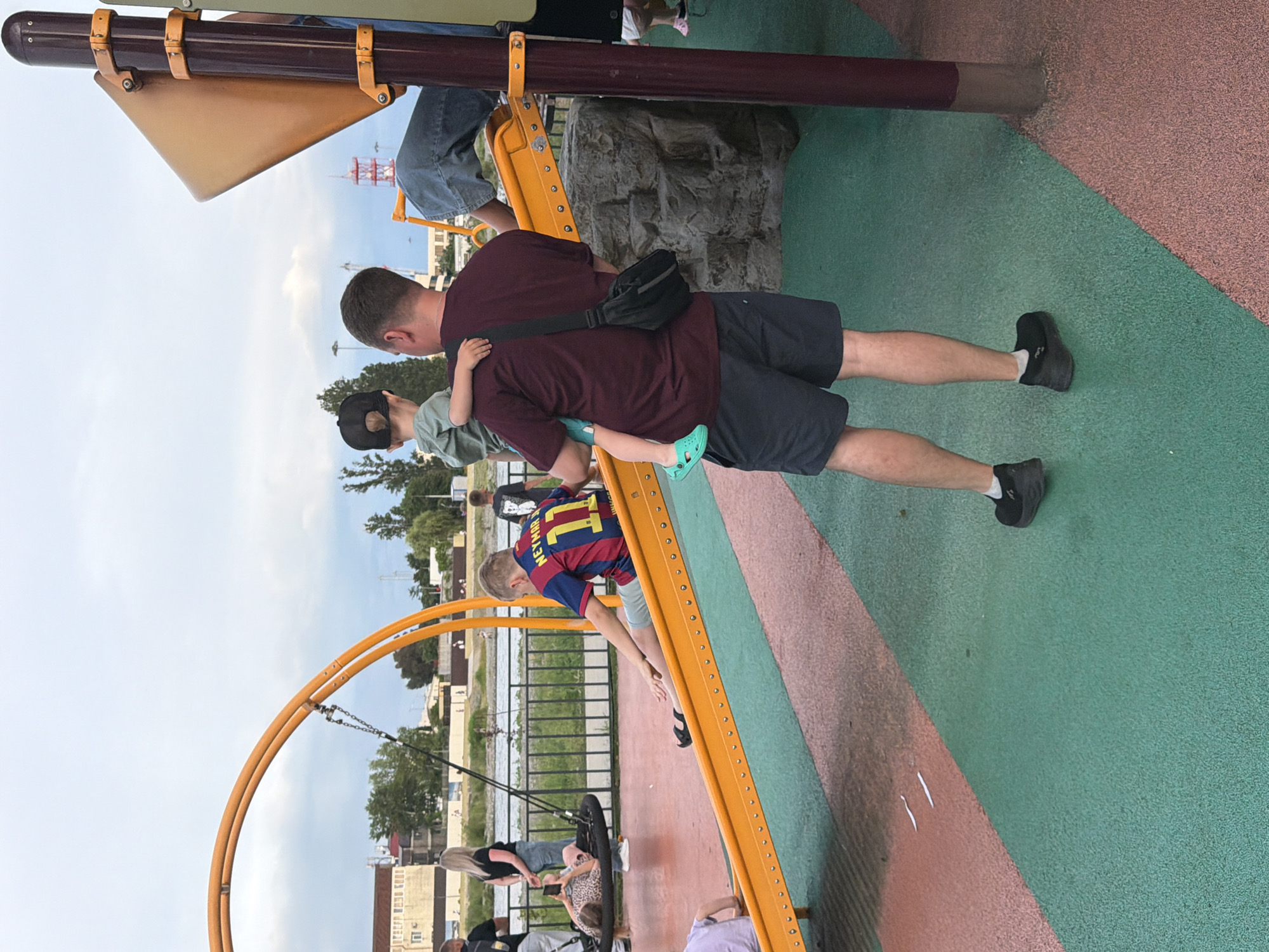 The image shows a playground scene in Sochi, Russia, on a cloudy day. A man is carrying a young child on his shoulders, facing away from the camera. The man is wearing a dark maroon t-shirt, dark shorts, and black shoes with white socks. The child is wearing a light green t-shirt and a black baseball cap, also facing away. To the left, a boy in a blue and red soccer jersey with the number 11 is sitting on an orange slide. He is wearing blue shorts and black sandals. Further left, a woman with blonde hair is standing near a circular swing set, where another child in a patterned dress is seated. In the background, there is a body of water with a fence along its edge, and buildings and trees are visible. The playground surface is a mix of red and green rubberized material. A large orange structure, part of the playground equipment, is prominent in the foreground and midground.