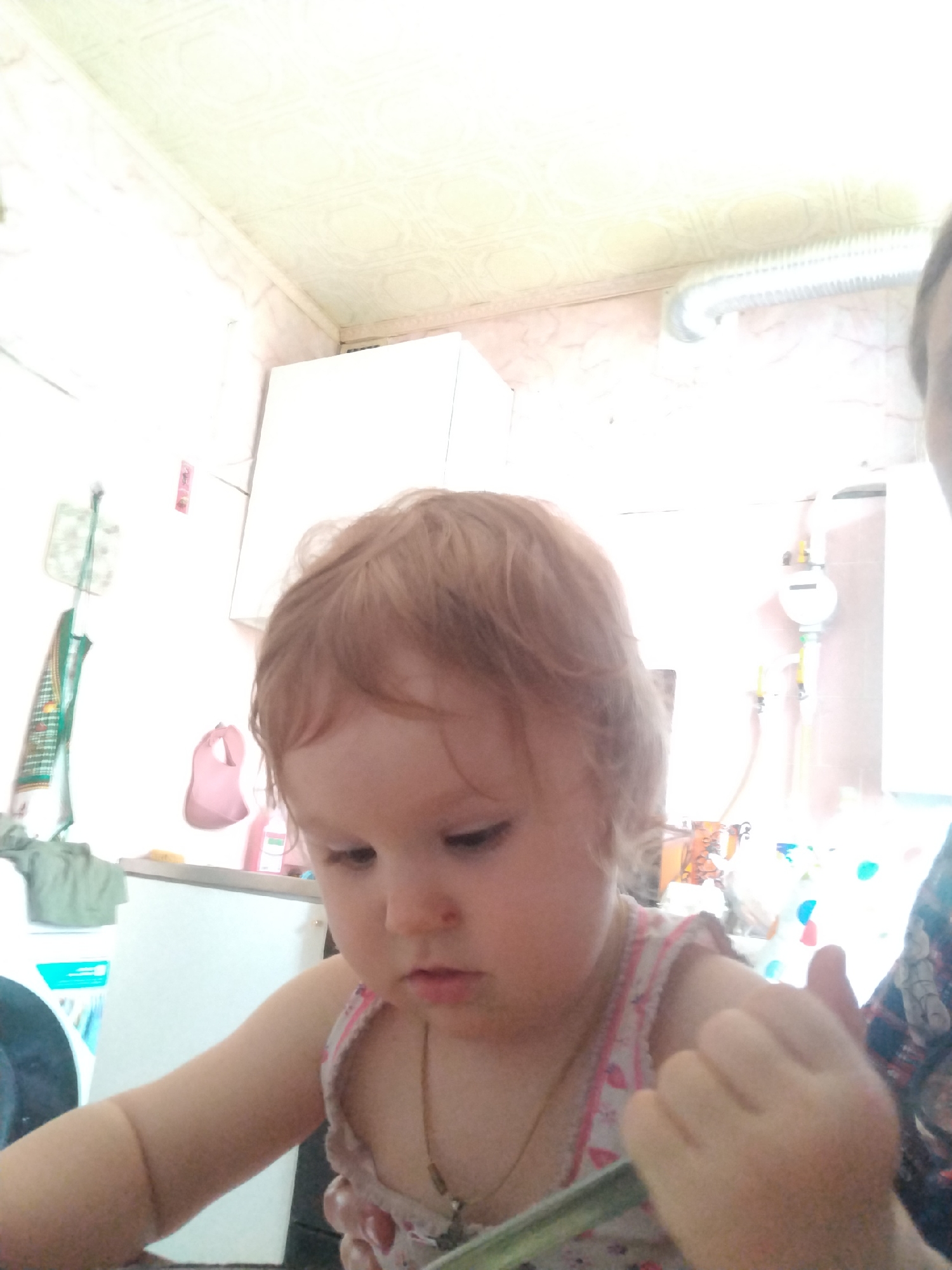A slightly blurred, frontal medium shot captures a young child, possibly a toddler, in an indoor setting that appears to be a kitchen or utility area in a residential building in Kaluga, Russia. The child, with light brown hair and fair skin, is looking down, seemingly engaged with something out of frame. They are wearing a light-colored garment with a patterned neckline.
The background reveals typical domestic features. To the left, a white appliance, possibly a refrigerator or washing machine, is partially visible. Above it, a green dishtowel or similar item hangs from a hook. A pink bib is also visible hanging on the wall. Further back, a white panel, possibly a cabinet or appliance, occupies a significant portion of the wall space. To the right of the child, a silver corrugated pipe extends horizontally from the wall, suggesting a ventilation system. Another white unit, possibly a water meter or boiler, with yellow and gray pipes, is mounted on the wall in the upper right quadrant. The ceiling has a textured, patterned finish. The overall lighting suggests artificial light from within the room. No other individuals are clearly identifiable besides the child, though a hand is visible in the lower right corner, possibly belonging to an adult.