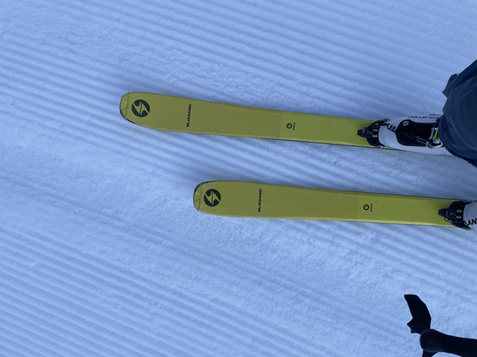 A first-person view depicts a skier's bright yellow Blizzard Zero G skis and Salomon ski boots on a meticulously groomed, ridged snow slope in Vallorcine, France. The skis are parallel, with bindings securing the boots. A partial black ski pole grip is visible in the lower-left, and the skier's dark blue pant leg is seen at the bottom center. The snow is pristine, showing distinct parallel corduroy patterns indicative of recent grooming.