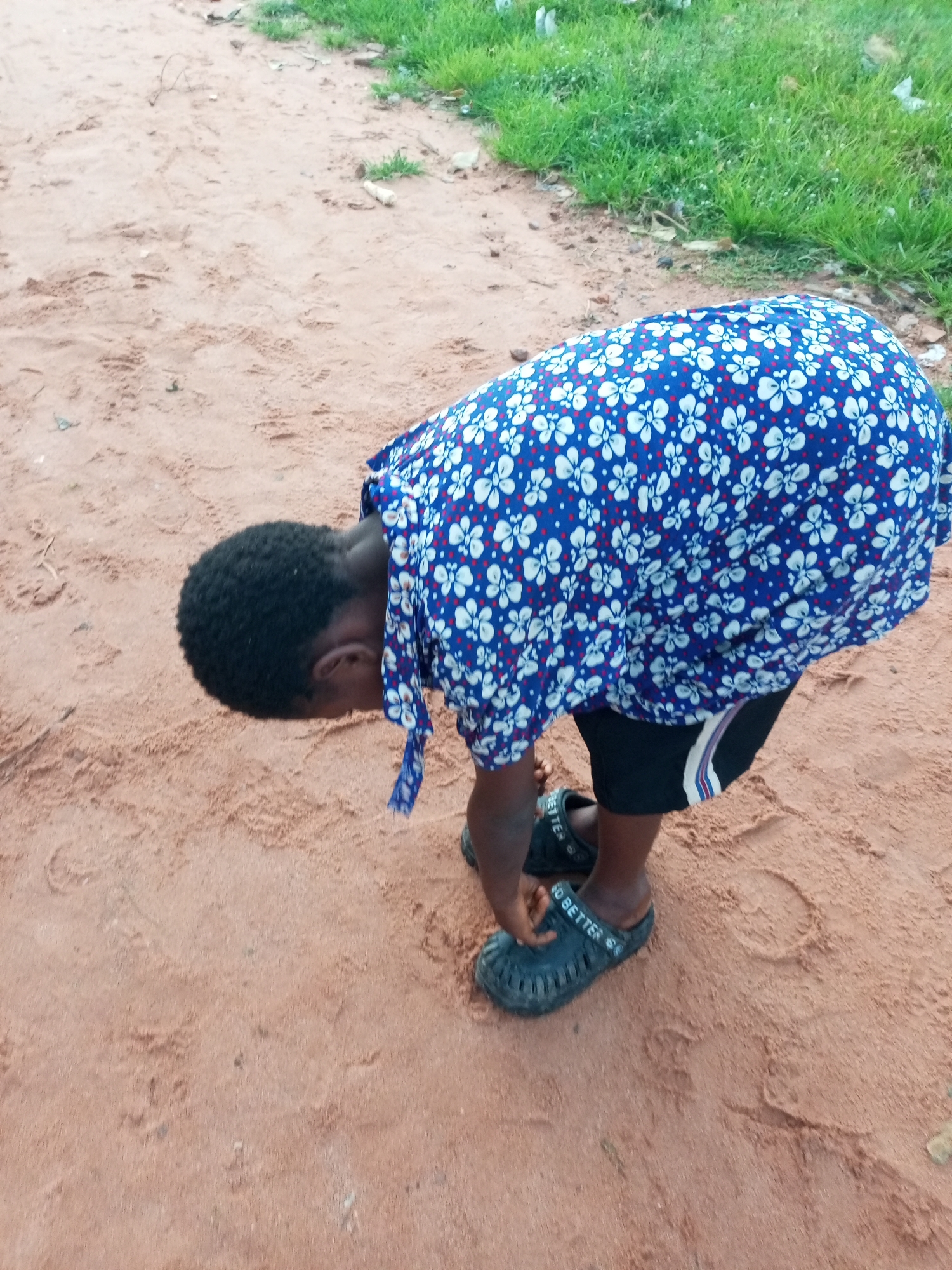 A young person is bent over on a dirt path in the city of Tempure, Nigeria, with their head down. They are wearing a blue dress with a white floral pattern and black shorts with white and red stripes. Their feet are clad in black sandals with the words "GO BETTER" printed on them. The ground is a reddish-brown dirt with some scattered grass and debris. The lighting suggests it is daytime.