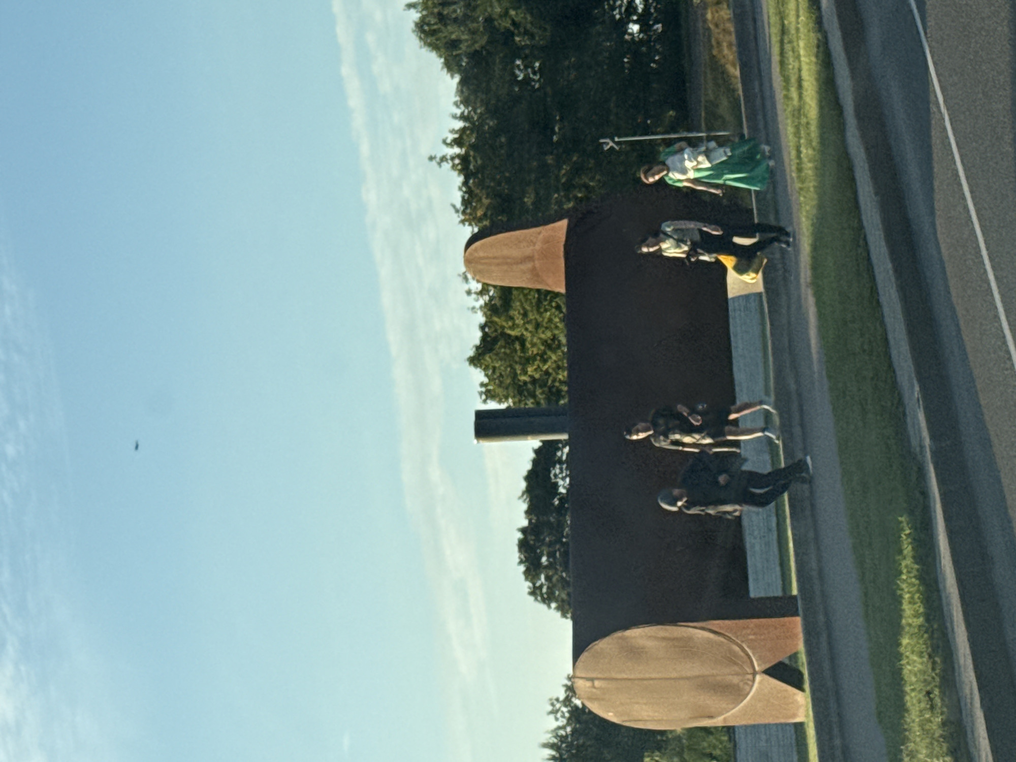 In Tallinn, Estonia, an outdoor scene unfolds under a clear, light blue sky with sparse white clouds. A large, dark, cylindrical sculpture, possibly a silo or a tank, dominates the mid-ground. It has a rounded end on the left and a peaked structure on the right. Four individuals are present near the sculpture. On the far left, a person wearing dark clothing, including a hooded jacket and dark pants, walks with their head down. Next to them, to the right, is another person in dark clothing, with short hair, walking. Further to the right, a person is wearing a light-colored top with a pattern and dark pants, carrying a dark bag with a yellow item. On the far right, a person in a long, bright green dress and a white garment draped over their arm walks. A faint white pole is visible behind the individuals on the right. The sculpture is situated on a grassy area next to a paved path or road, with more trees visible in the background. A small, dark object, possibly an airplane, is visible in the sky on the upper left of the frame. The ground in the foreground is paved, with grassy verges.