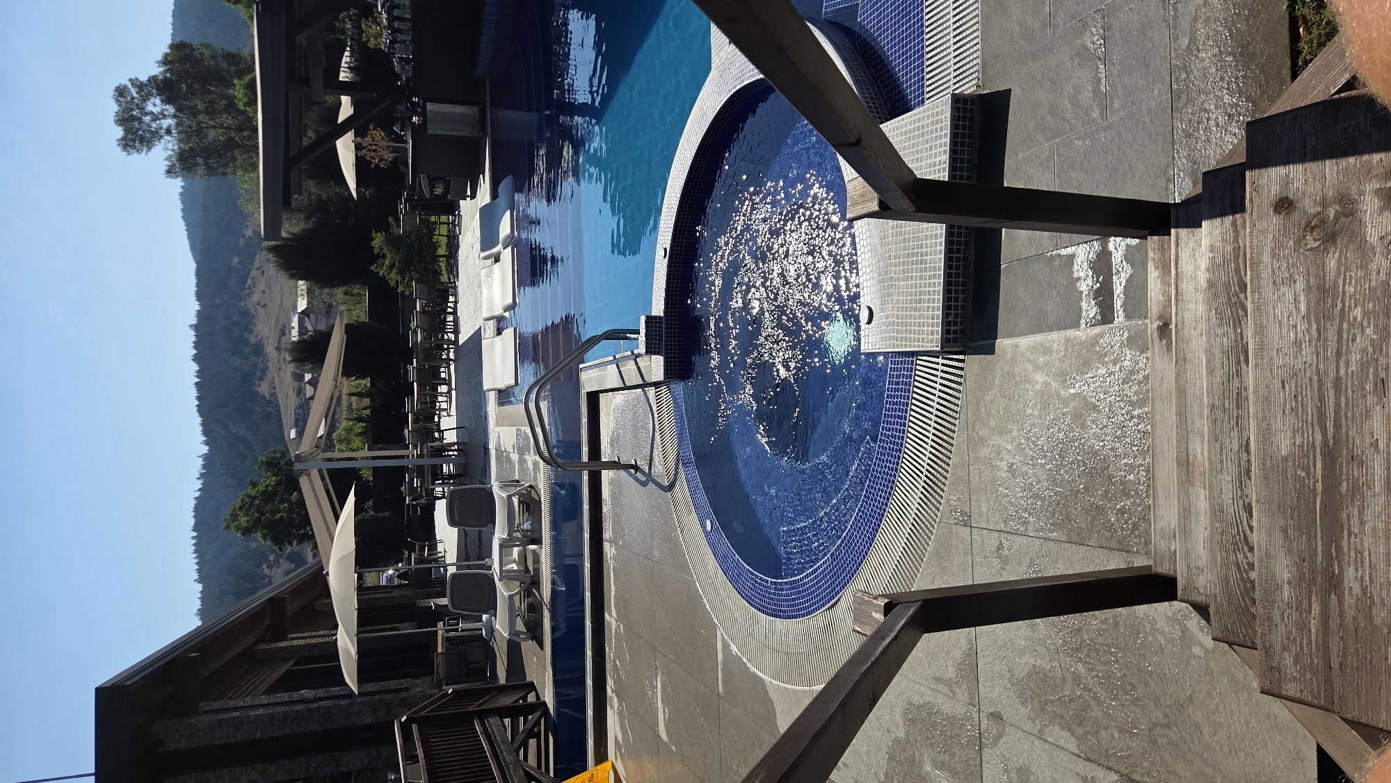 The image shows an outdoor swimming pool area, likely a resort or hotel, in the city of Bania, Bulgaria. In the foreground, a set of weathered wooden stairs with dark wooden railings leads down towards the pool area. The stairs appear to be wet, with visible water droplets and streaks on the surface. To the left and right of the stairs, a grey tiled patio area is also wet, indicating recent activity or cleaning.
Dominating the mid-ground is a circular, tiled hot tub, filled with bubbling blue water that reflects the bright sunlight. The hot tub is surrounded by a tiled edge and a drainage grate. Beyond the hot tub, a larger swimming pool with clear blue water stretches into the background. Several white lounge chairs are visible near the edge of the larger pool, some partially submerged.
In the background, a building with a stone facade and a wooden roof structure can be seen to the left. Umbrellas provide shade over outdoor seating areas with tables and chairs. Further back, a lush green hillside covered with trees forms the backdrop, with some buildings nestled amongst the foliage. The sky is a clear, bright blue, suggesting a sunny day. No people are clearly visible in the image, though the wet surfaces and lounge chairs suggest recent occupancy.