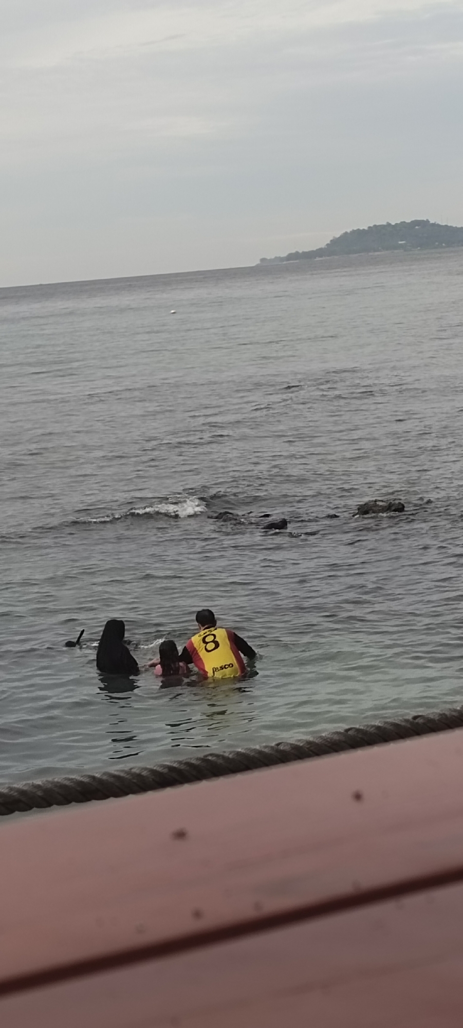 A group of three people, two adults and one child, are standing in shallow water. One adult, wearing a black hijab, is on the left. A child, wearing a pink swimsuit, is in the middle. Another adult, wearing a yellow and red shirt with the number "8" and "PASCO" written on it, is on the right, facing the child. The water is murky and there are waves and rocks visible. In the background, a hilly island with trees and buildings is visible on the horizon. The sky is overcast and grey. The foreground shows a wooden structure with a thick rope. The scene appears to be at a beach or waterfront location in Sabang, Indonesia.