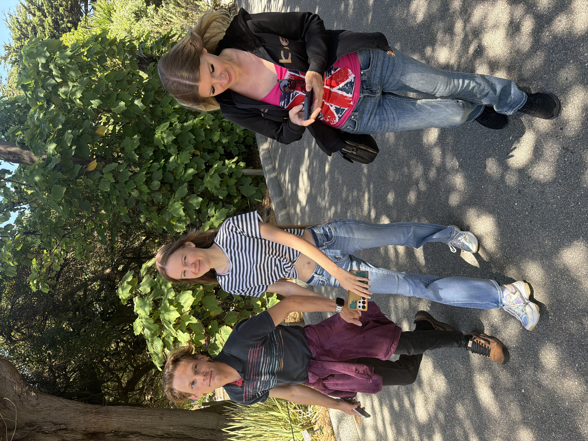 Three people are walking on an asphalt path outdoors. The person on the left is a man wearing a dark polo shirt with horizontal stripes, black pants, and brown sneakers. He is holding a cell phone in his left hand and has a maroon jacket tied around his waist. Next to him is a woman wearing a horizontally striped t-shirt tied at the waist, blue jeans, and white sneakers. She is holding a cell phone in her right hand. On the right is another woman wearing a black hooded jacket over a pink tank top with the Union Jack design, blue jeans, and black sneakers. She is looking at her cell phone, which she holds in her right hand. All three individuals are looking in different directions. The background consists of lush green trees and vegetation, with dappled sunlight filtering through the leaves, creating shadows on the path. The scene appears to be a park or a scenic trail. Given the presence of cypress trees, the vegetation, and the clear sunny weather, this scene is consistent with areas like Golden Gate Park or the Presidio in San Francisco, California, United States of America.