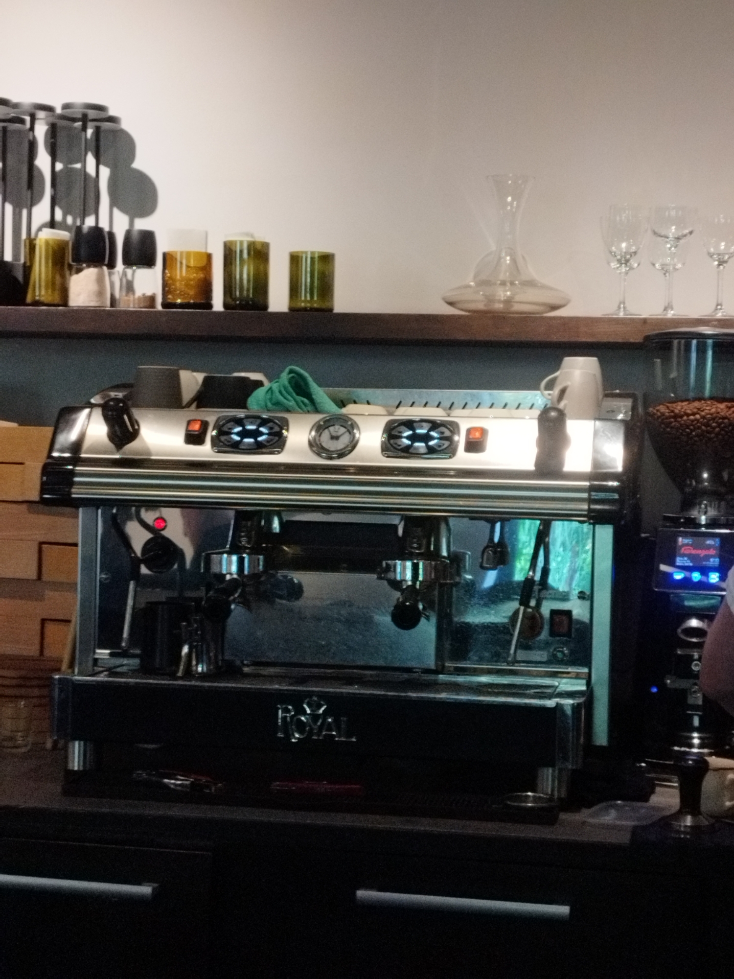 The image depicts an espresso machine and various accessories on a shelf and counter in what appears to be a coffee shop in Saint Petersburg, Russia. On the top shelf, several glass containers of different colors and sizes are visible, along with a decanter and wine glasses. Below the shelf, the espresso machine is the central focus. It's a silver and black machine with various buttons, levers, and a portafilter. To the right of the espresso machine, a coffee grinder filled with beans is positioned. The scene suggests a working coffee establishment, with the equipment ready for use. The overall lighting is ambient, contributing to a relaxed atmosphere.