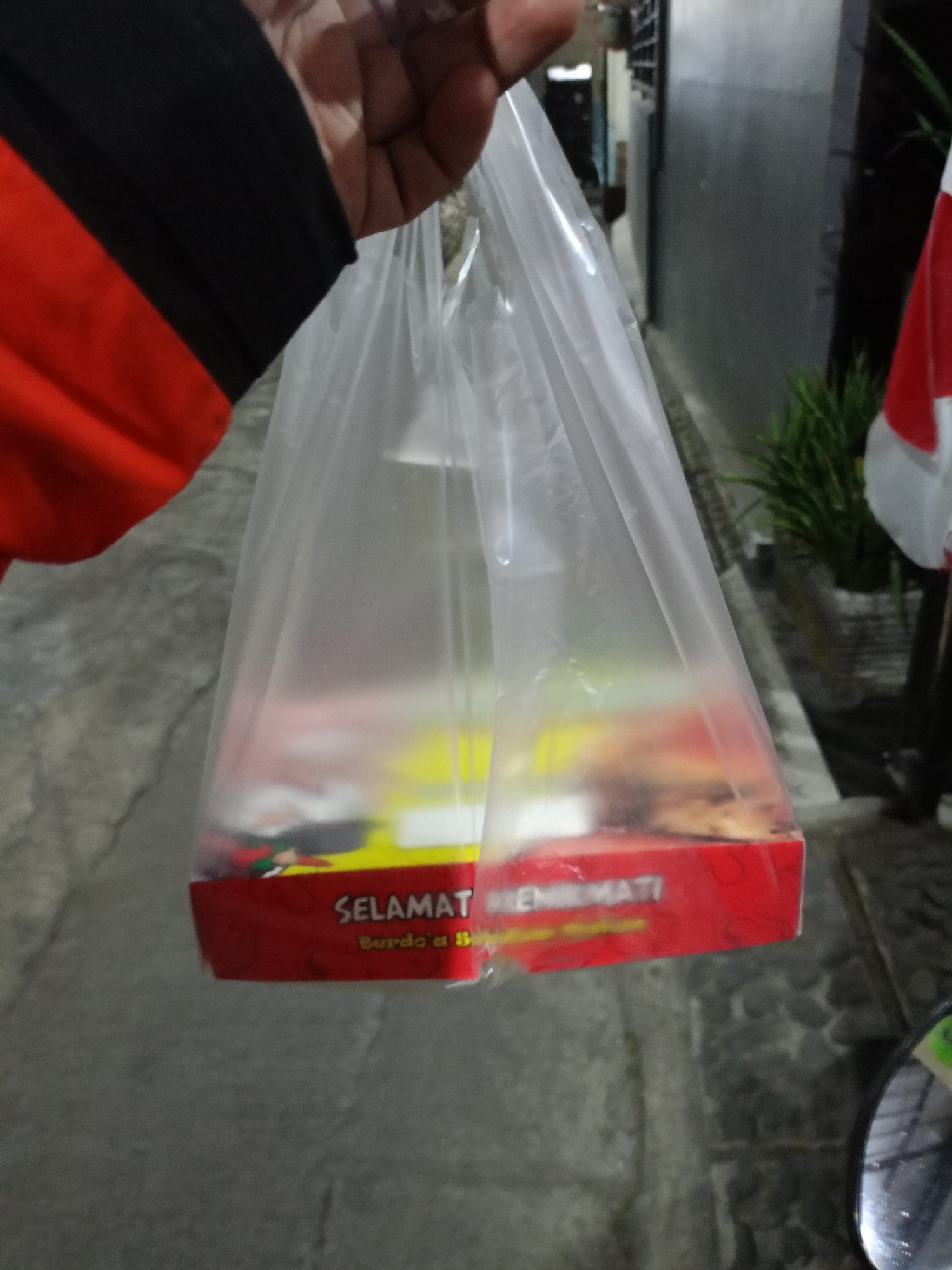 A person's hand, wearing a red and black jacket, holds a clear plastic bag containing food. The bag is opaque due to the contents inside. The bottom of the food packaging is visible, showing a red and yellow design with text that appears to read "SELAMAT MENIKMATI" and "Burdo a B". The background is blurred, but suggests an outdoor urban environment, possibly a street in Batu, Indonesia, with a textured gray ground and a dark building to the right. A potted plant and part of an Indonesian flag are also visible in the background. A motorcycle mirror is visible in the lower right corner of the image.
