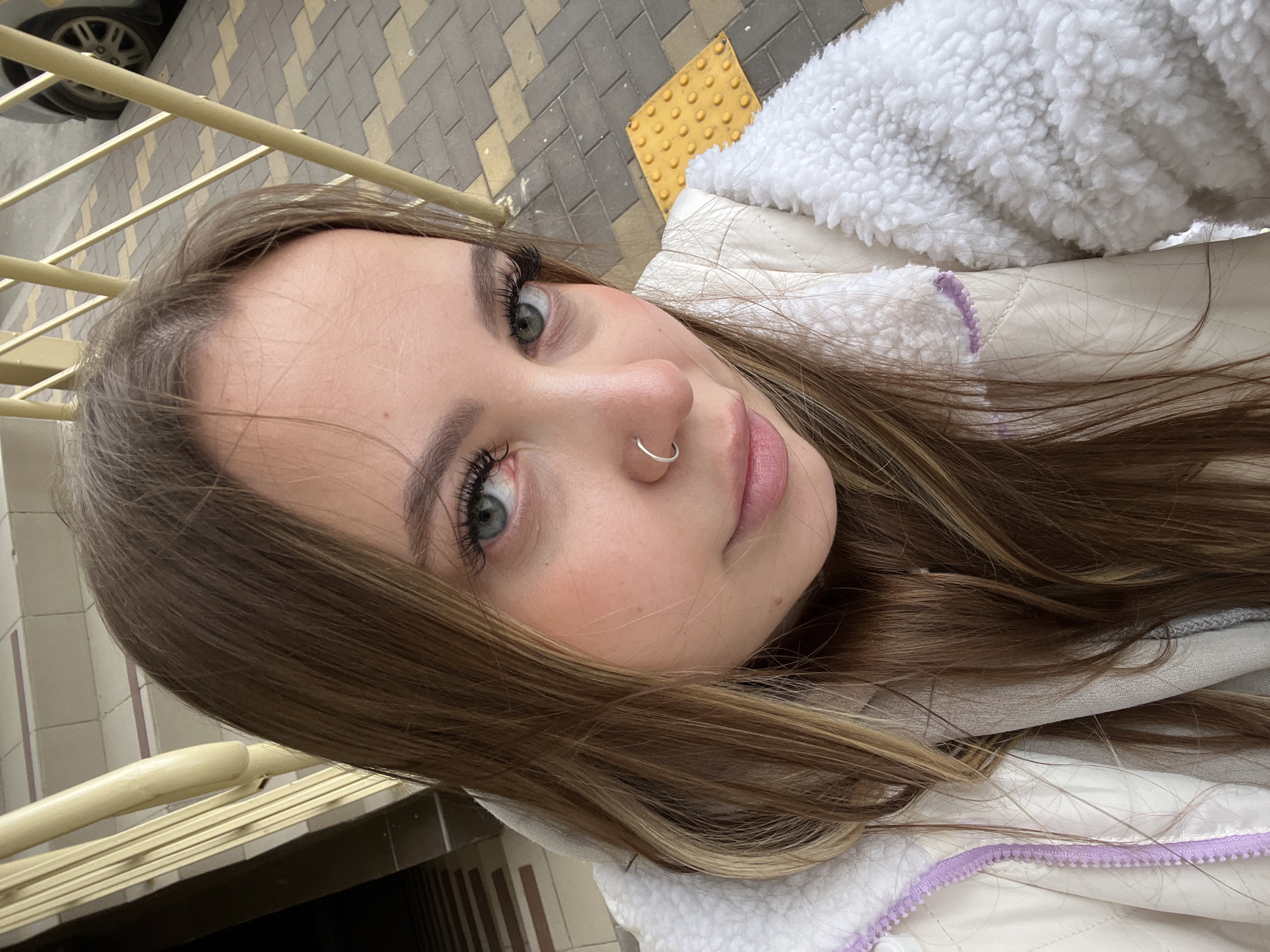 A young woman with light brown hair and light-colored eyes is captured in a close-up, high-angle selfie. She has long, dark eyelashes, light pink lips slightly parted, and a silver nose ring in her left nostril. Her hair is parted and falls around her face and shoulders.
She is wearing a cream or white jacket with a prominent white fluffy, sherpa-like lining, visible around the collar and edges. A hint of a light purple zipper or trim can be seen on her jacket.
The background suggests an outdoor urban setting during daytime, with ample natural light. To the upper left, a car wheel is partially visible on what appears to be a paved street. Below this, gray and beige paving stones are laid in a pattern, alongside a yellow tactile paving slab with raised dots, typically used for the visually impaired. A light-colored metal railing runs diagonally across the image, suggesting the woman is positioned near a staircase, balcony, or elevated walkway. Her expression is neutral and direct, looking towards the camera. The jacket implies a cool weather setting, possibly characteristic of Krasnodar, Russia. No visible text is present in the image.