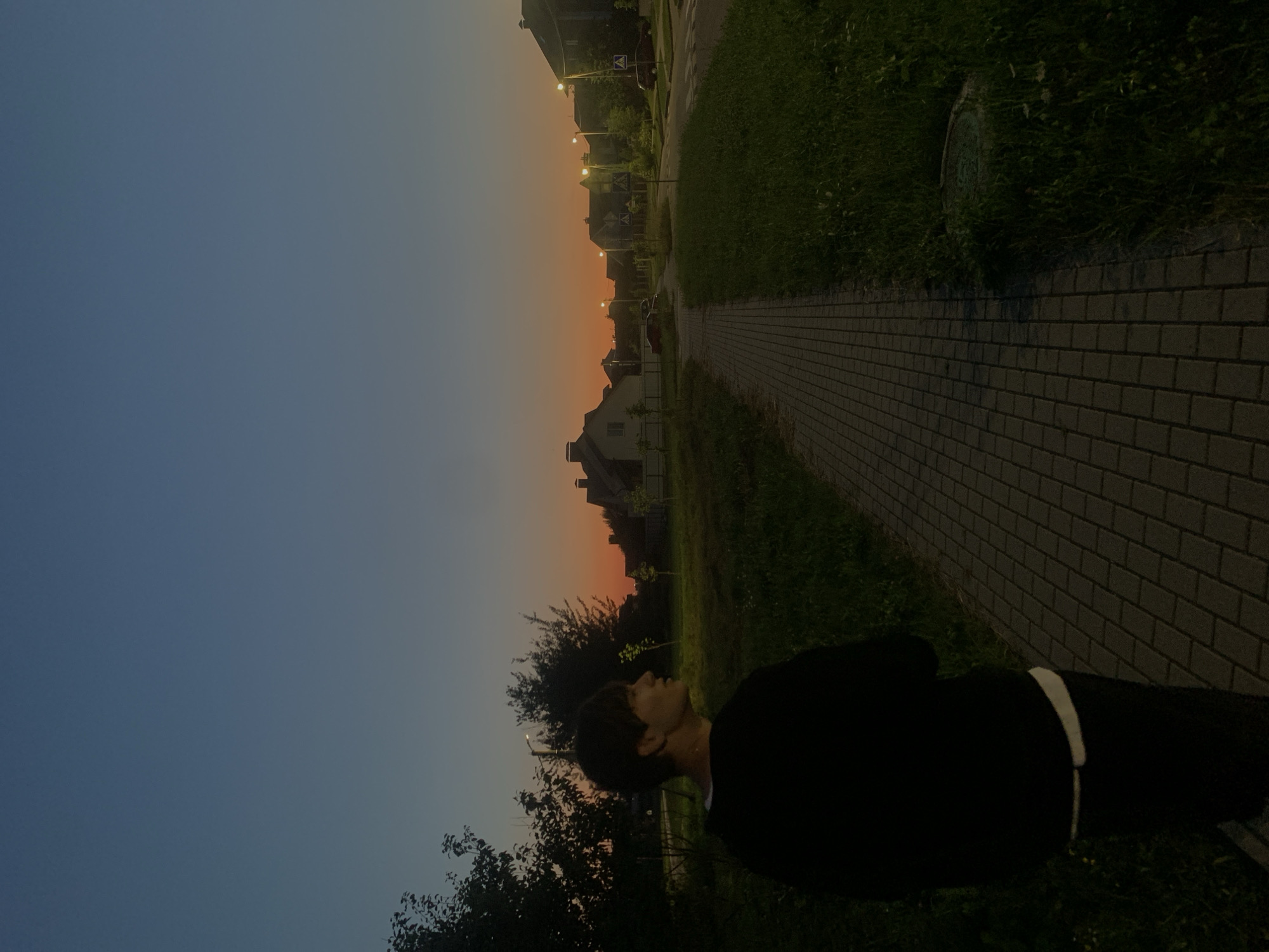 A lone figure, facing away from the camera, walks along a brick pathway at dusk. The person is wearing a dark long-sleeved shirt and dark pants with a white waistband visible. The pathway is bordered by green grass on both sides. In the background, a street with houses and streetlights can be seen. The sky transitions from a deep blue overhead to a warm orange and yellow gradient near the horizon, indicating either sunrise or sunset. This scene is consistent with a suburban residential area in Minsk, Belarus, during twilight.
