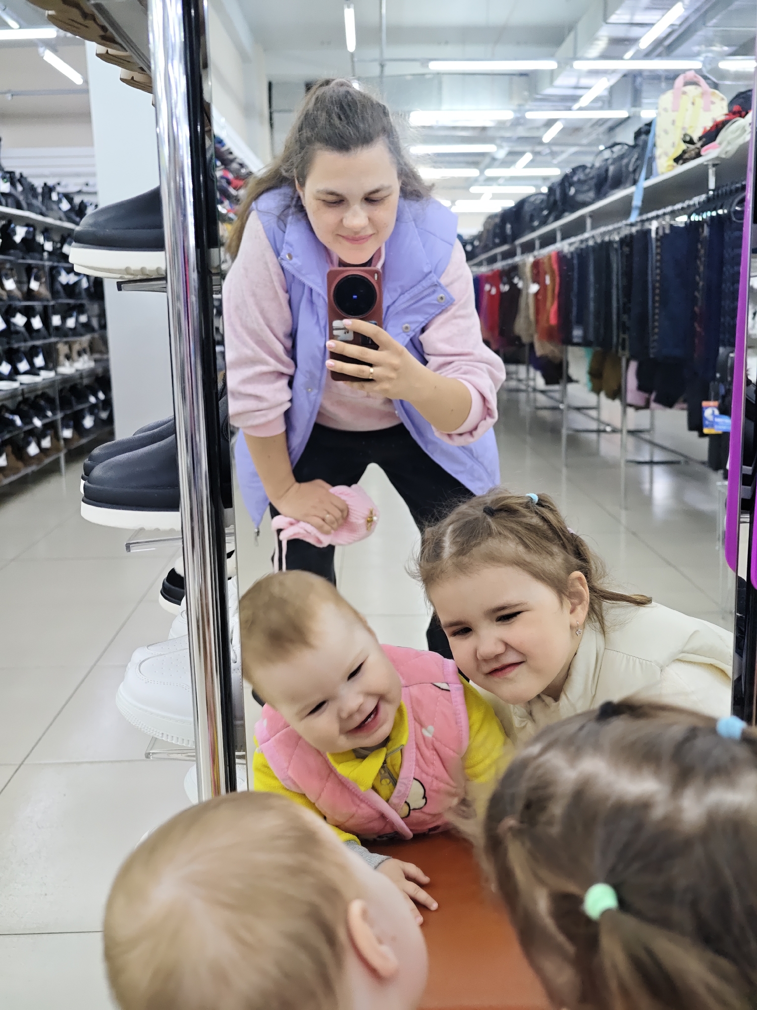 A woman is taking a selfie in a department store mirror with two young children in the city of Chaikovsky, Russia. The woman is wearing a pink sweater, a lilac vest, and black pants, and has her hair pulled back. She is holding a red-brown phone and looking at the screen. In the mirror’s reflection, two young girls are visible. One, wearing a yellow top and pink vest, is smiling towards a baby on the other side of the mirror. The second girl, wearing a cream-colored jacket, is looking toward the baby. Part of a third child's head is visible at the bottom of the mirror. The background shows shelves stocked with shoes and rows of clothing racks. The floor is white tile. Overhead fluorescent lights are visible.