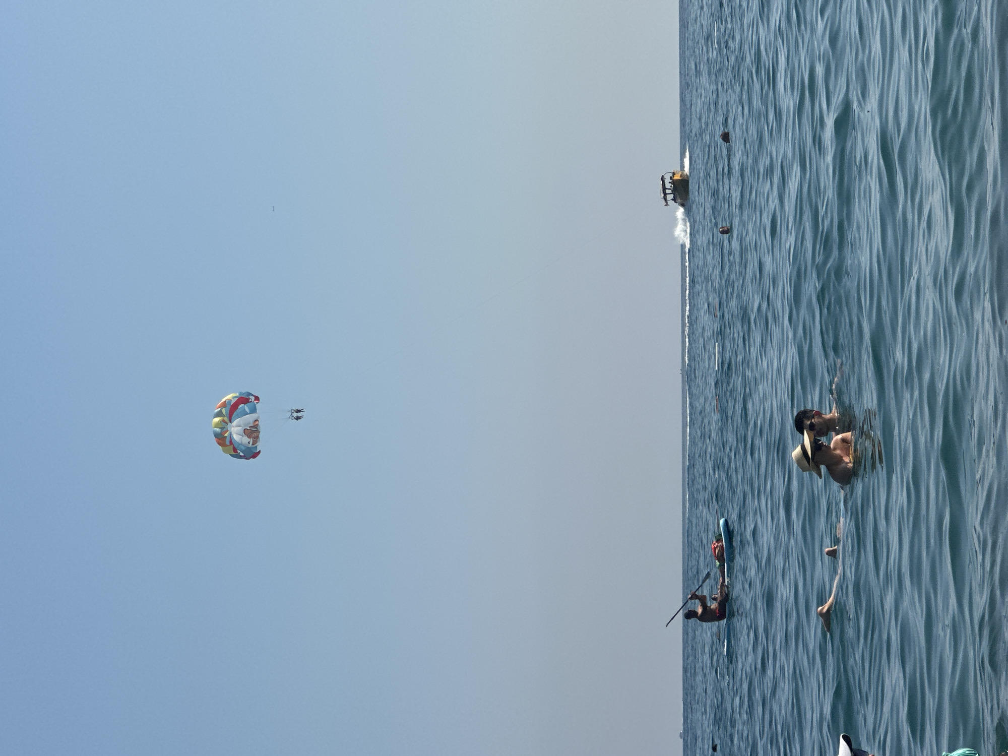 In Boreti, Montenegro, on a clear day, a parasail with a colorful canopy and a cartoon character motif is visible in the sky, carrying two individuals. Below, on the sea, a person is paddleboarding. Further in the distance, a yellow motorboat is speeding across the water, leaving a wake. Closer to the foreground, two people are swimming, one wearing a straw hat and sunglasses, with another person beside them. The sea is a deep blue with moderate waves.