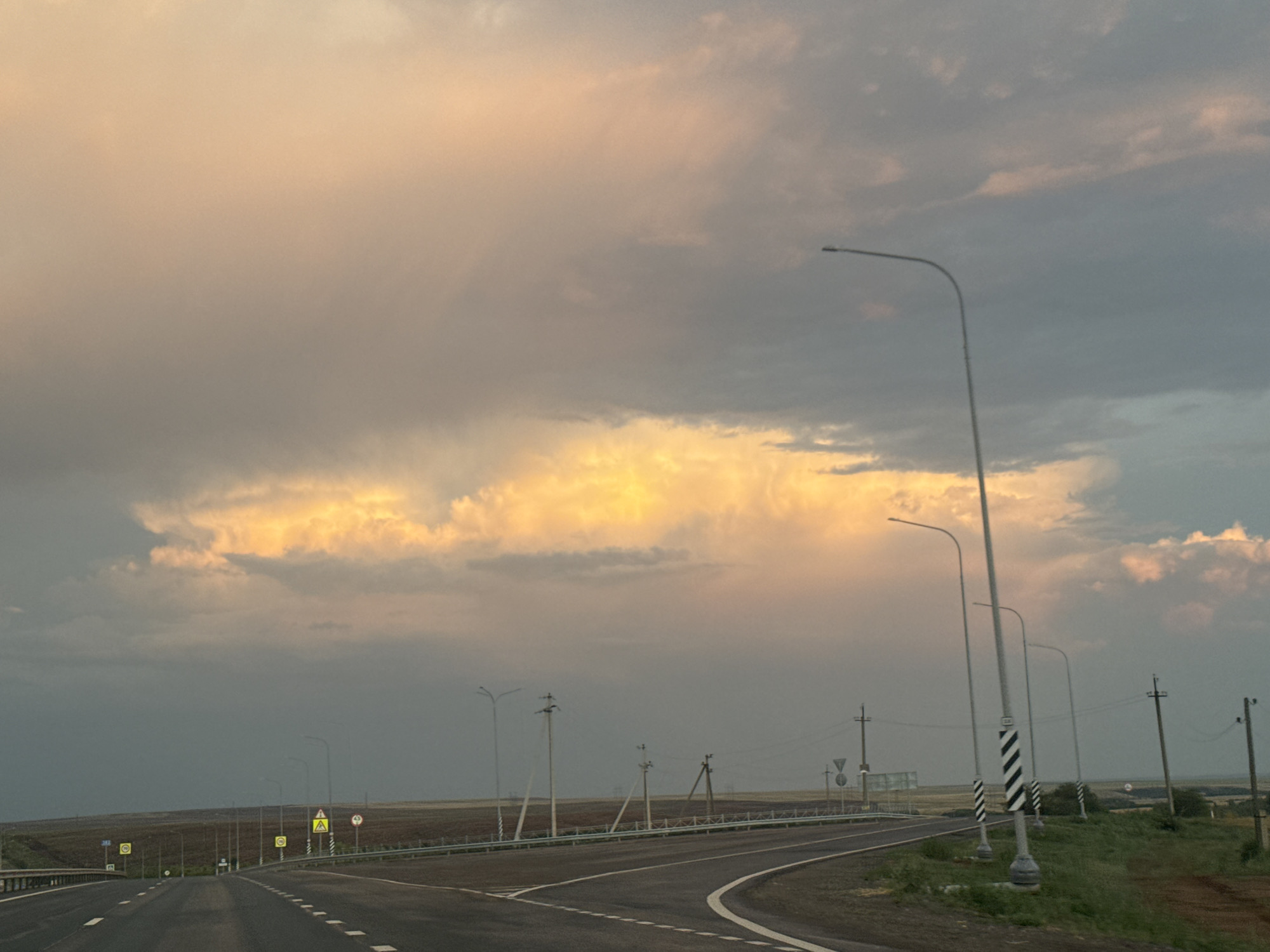 The image depicts a highway landscape under a dramatic sky, likely in the vicinity of Syrt, Russia, given the context. The sky is dominated by heavy clouds, with a bright band of golden light piercing through them horizontally, suggesting either sunrise or sunset. The road ahead curves to the right and is divided by white lines. Several streetlights line the road, some with diagonal black and white markings. Various road signs are visible, including cautionary signs with yellow backgrounds and circular signs. The surrounding environment appears to be agricultural land, with fields visible beyond the road. No people or vehicles are actively depicted in motion, contributing to a sense of stillness.