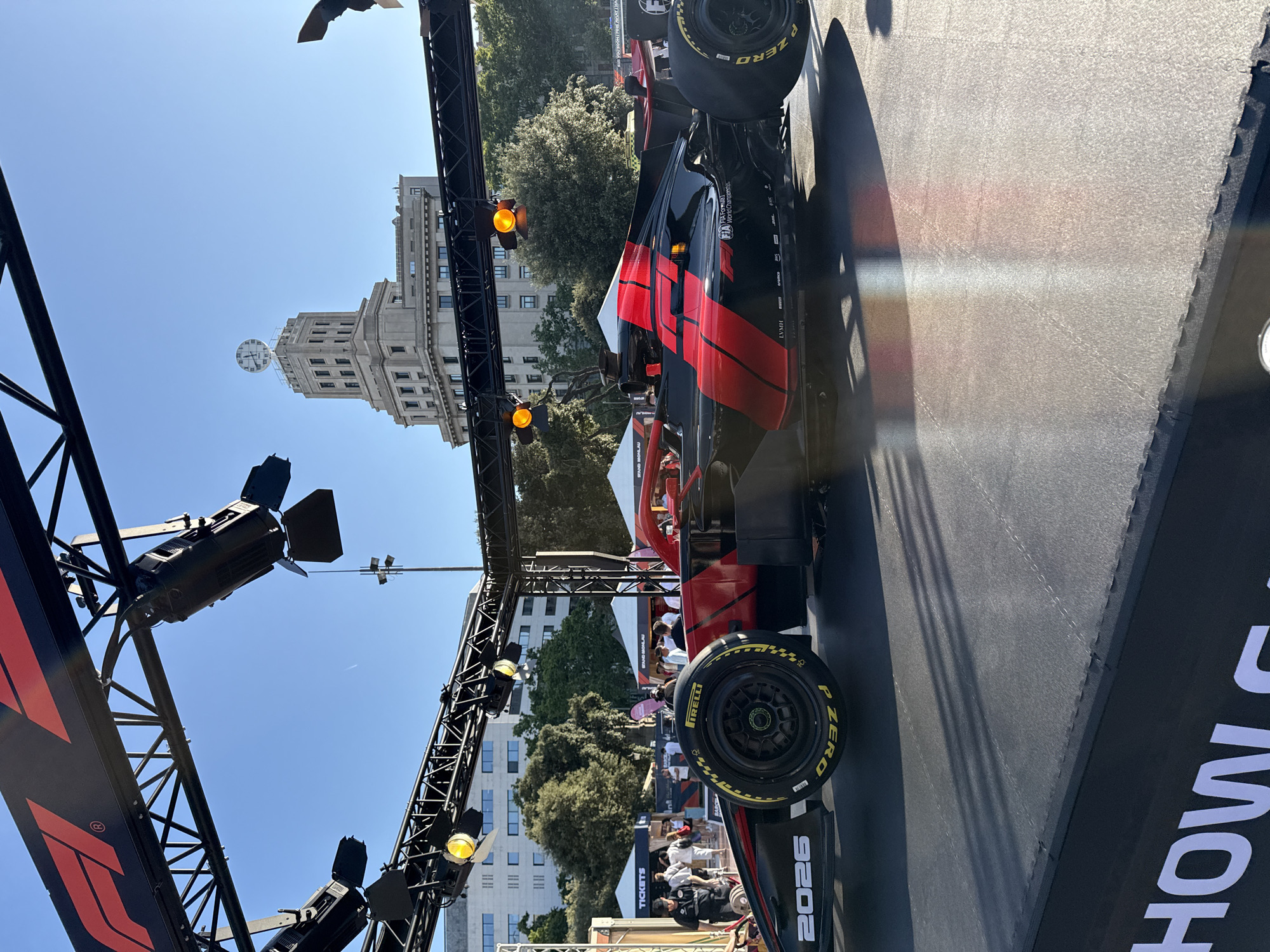 The image shows a black and red Formula 1 car parked on a black platform in front of a building with a clock tower, located in Barcelona, Spain. The Formula 1 logo is visible on a black truss structure above the car, which is illuminated by spotlights. The car has the number "2026" printed on its side and the Pirelli P Zero tires are visible. In the background, there are trees, tents, and people walking around. A banner with white text on a black background is visible at the bottom of the image. The sky is clear and blue.
