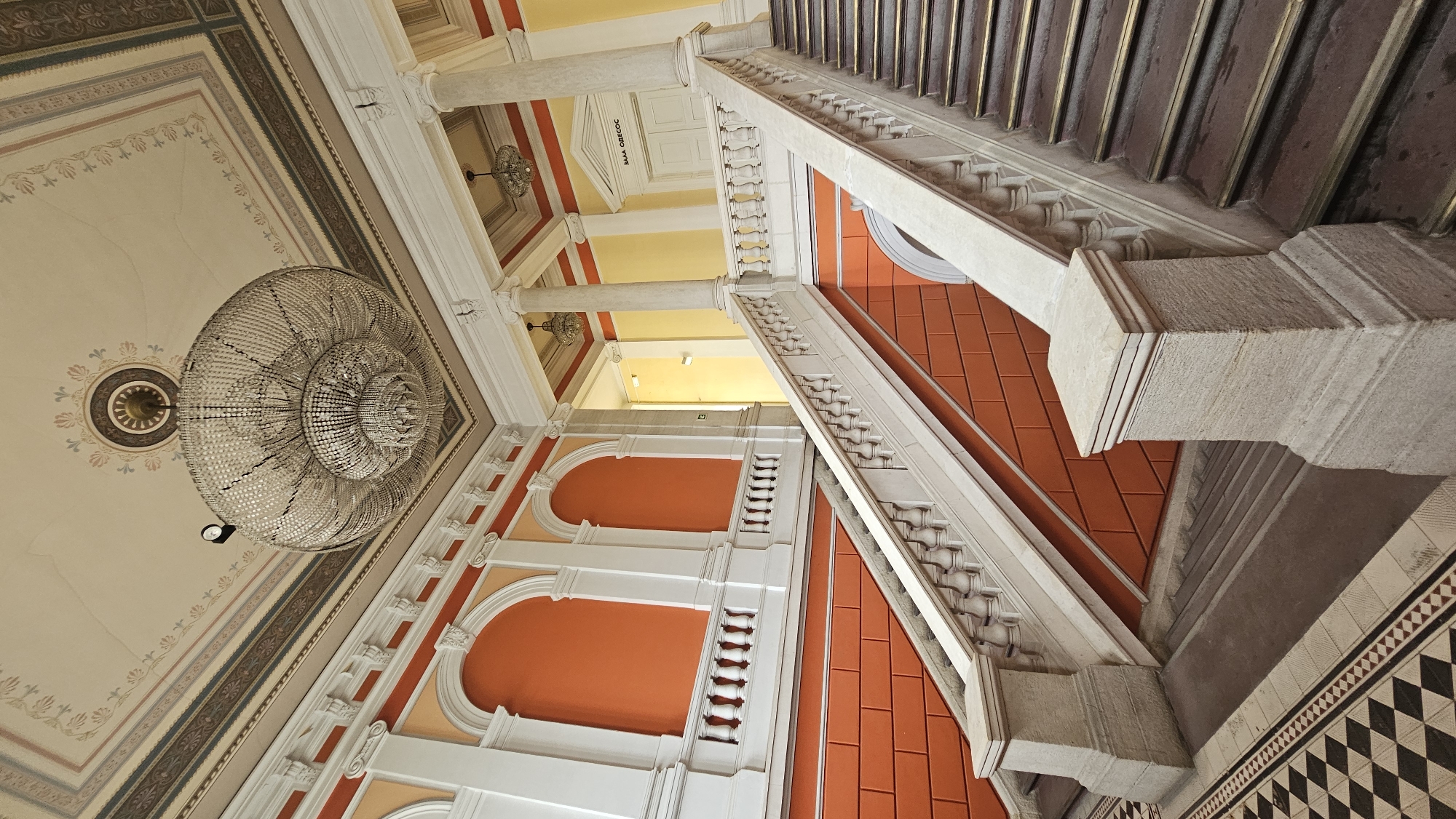 The image captures the interior of a building in Varna, Bulgaria, featuring a grand staircase and ornate architectural details. The perspective is from below, looking upwards. A large, spherical chandelier hangs from the ceiling, which is decorated with intricate patterns. Below, a series of columns support the upper levels, with visible arched openings and orange-painted walls. A prominent staircase with white balustrades and brown steps ascends to the right of the frame. The building's style suggests historical or cultural significance. A sign with "SALA OBRETS" is visible on a door above the pillars.