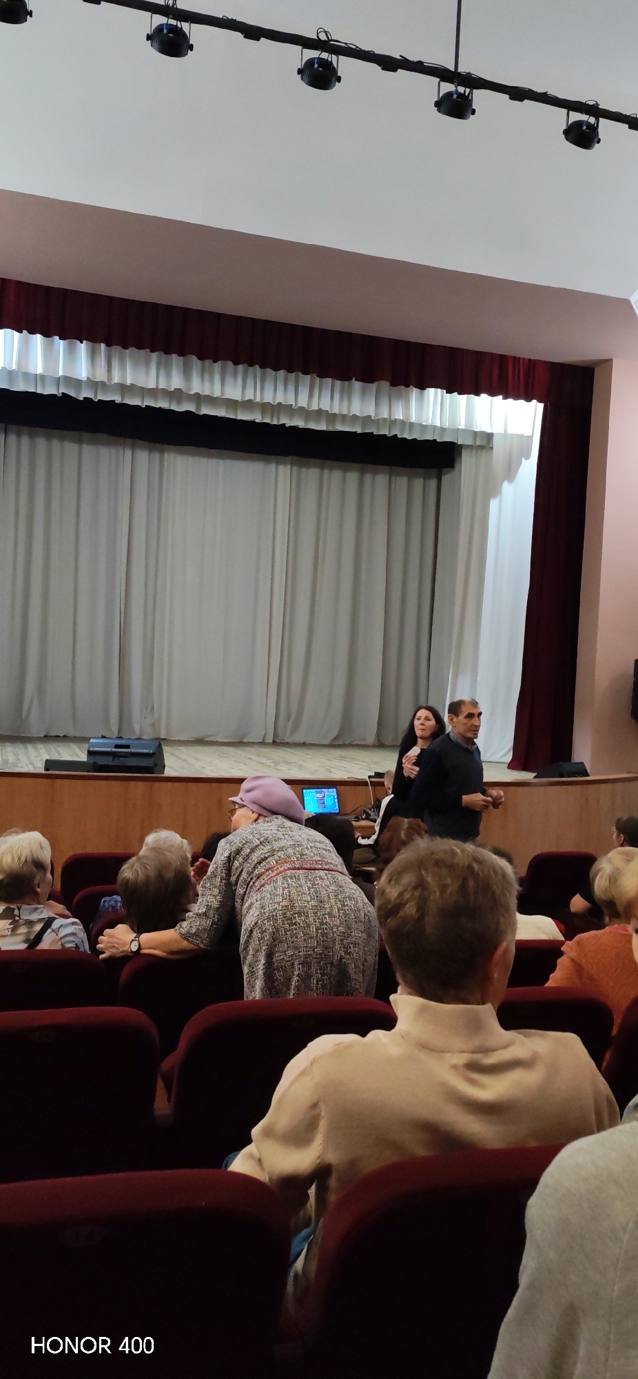The image shows the interior of a small theater or auditorium. There is a stage with white and maroon curtains. On the stage, a man and woman are standing. In the foreground, there are several rows of red upholstered chairs occupied by a mostly elderly audience. One woman in the audience is wearing a purple hat and grey dress. The photograph was taken in Noviy, Russia, as implied by the AI model name HONOR 400.