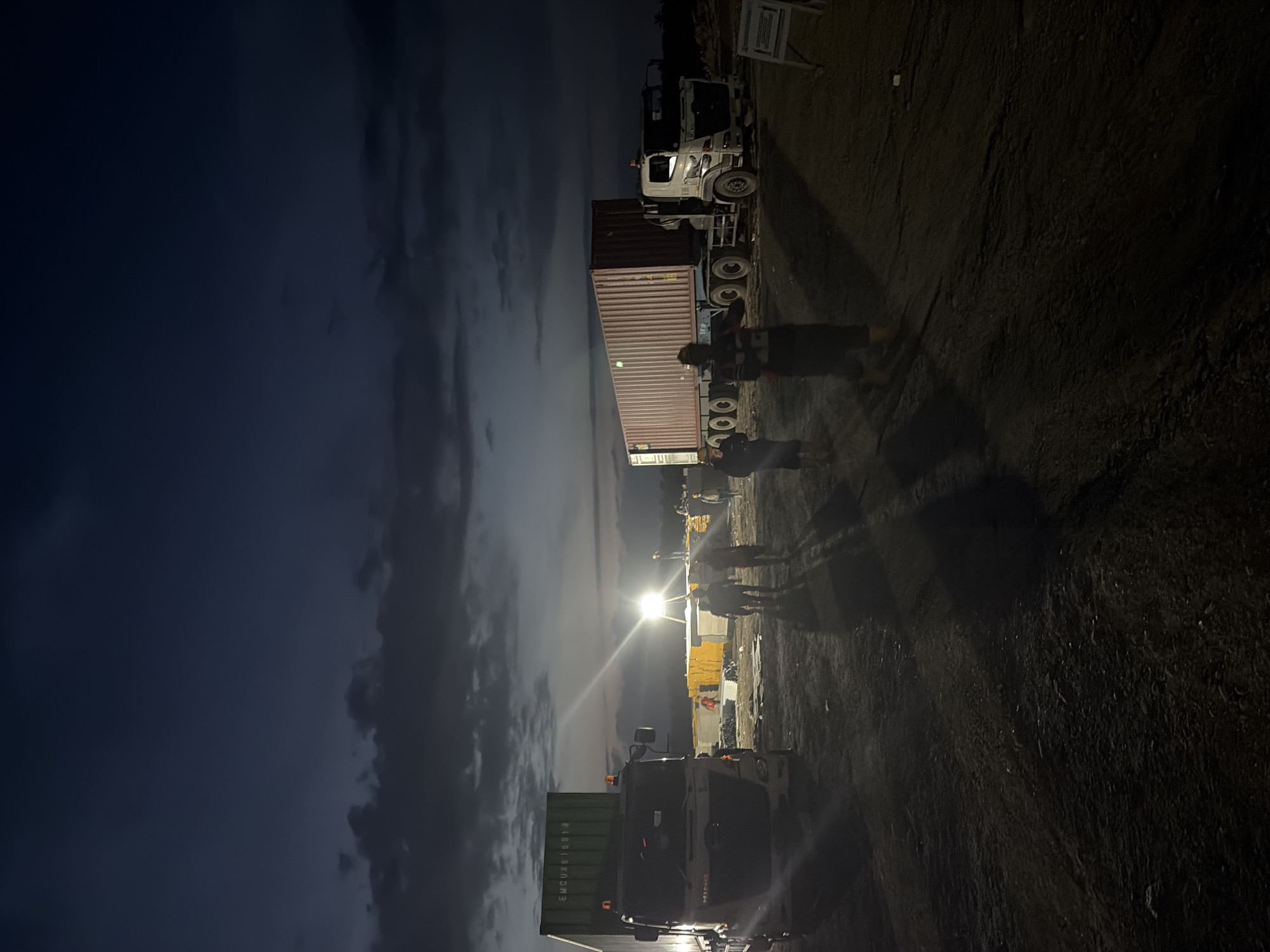 The image shows a nighttime scene in Simpangempat, Indonesia, featuring several trucks and people. The sky is cloudy. Two trucks carrying shipping containers are visible, one in the foreground with a green container and another further away with a red container. There is a group of people standing near the trucks, casting long shadows due to a bright light source in the background. The ground appears to be unpaved.