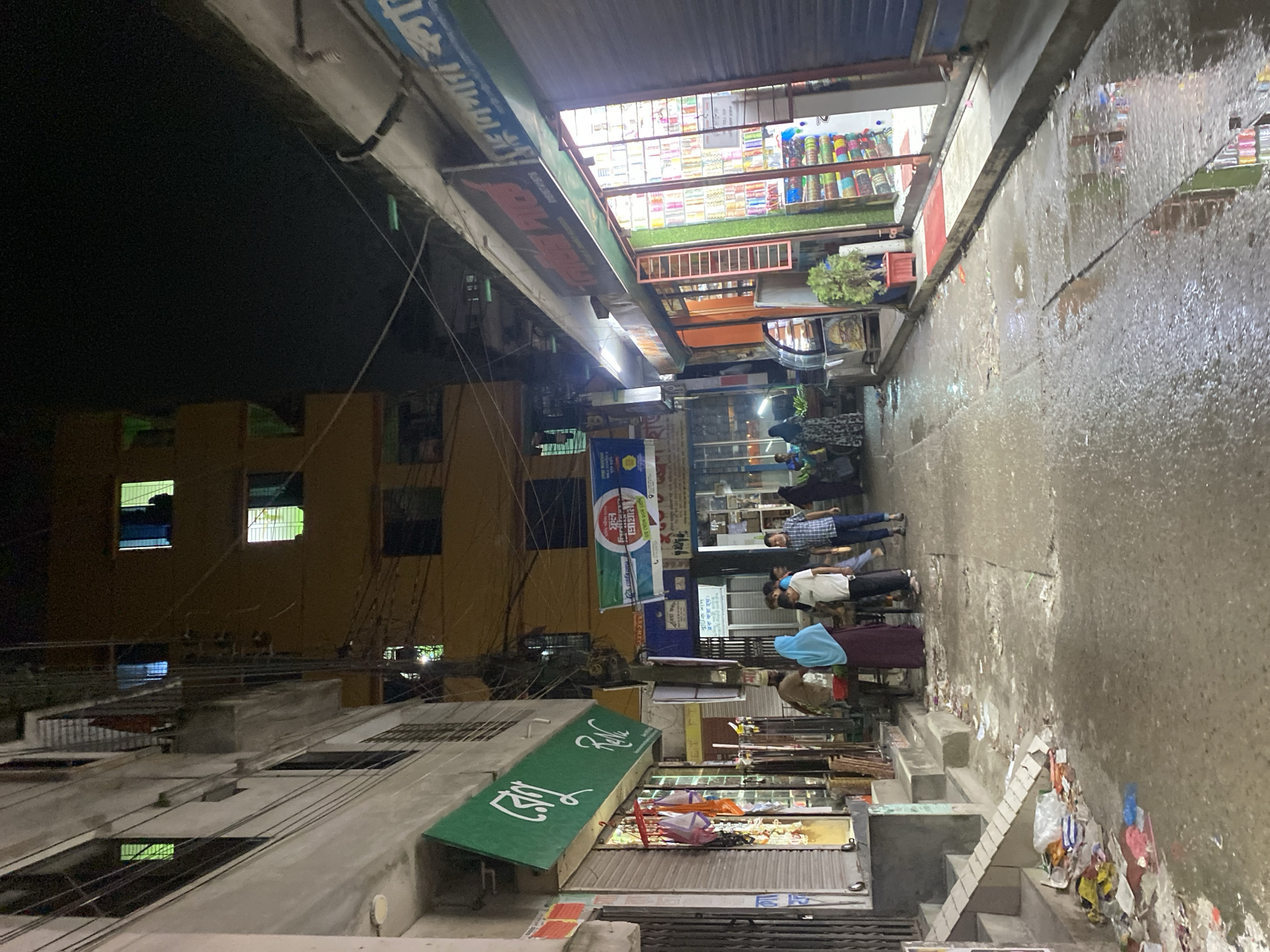 This is a nighttime street-level view of a narrow alleyway in Dhaka, Bangladesh, likely a commercial area. The scene is illuminated by artificial lights from shops and residential buildings, casting reflections on the wet, cluttered pavement. The alley is lined with multi-story buildings on both sides, featuring shops with their shutters down or partially open, displaying various goods.
On the left, a building shows a green awning with Bengali text, likely a shop name. Clothes or bags are hanging outside this establishment. Further down the alley, a group of people are gathered, including a woman in a blue headscarf and a dark dress, and a man in a brown shirt and a white cap. Another building on the left has several windows with illuminated interiors, one showing a person silhouetted against the light.
On the right, a brightly lit shop showcases shelves filled with colorful items, possibly textiles or bangles. Above this shop, a sign with Bengali script is visible. The alley floor is uneven, with visible cracks and puddles of water, strewn with litter such as plastic bags, paper, and other debris. Overhead, a complex network of electrical wires crisscrosses the sky. The overall impression is one of a bustling, though somewhat unkempt, urban environment at night.