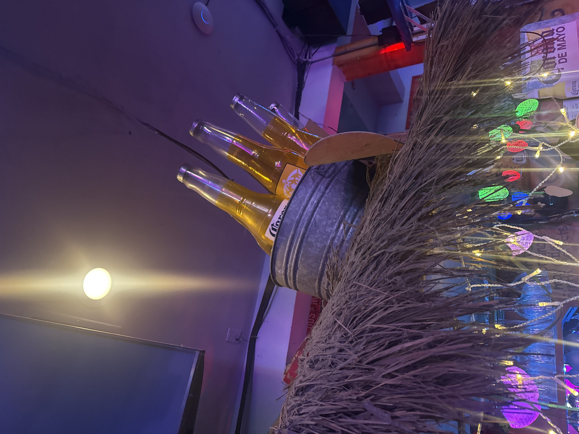 A low-angle shot looking up at a metal bucket filled with Corona beer bottles and surrounded by a thatch-like decoration, possibly taken in a bar or restaurant setting. The bottles are angled outwards from the bucket. Small decorative lights are intertwined with the thatch.
In the background, there is a ceiling with a circular light fixture and possibly some elements of a wall or part of the interior structure. The lighting in the scene is a mix of natural and artificial light, creating a somewhat stylized atmosphere. The colors are vivid and bright. The location is West Hollywood, United States of America.