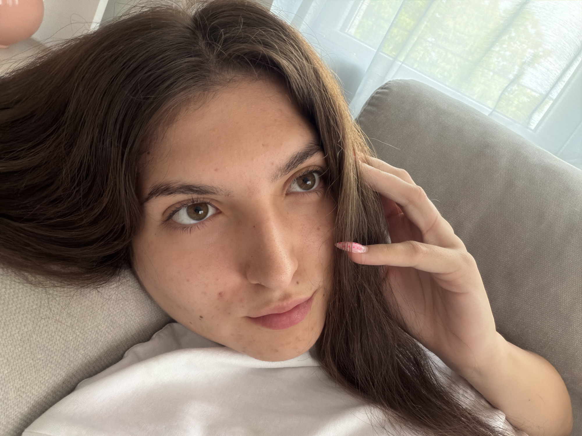 A young woman with long, dark brown hair is lying on a sofa. She is looking towards the camera with a neutral expression. Her right hand is raised to her face, touching her hair. She is wearing a white t-shirt. The background shows a window with sheer curtains and a portion of a grey sofa. The image appears to be a selfie taken indoors. There are no identifiable landmarks or details that would suggest a specific location within the city of Stara Zagora, Bulgaria.