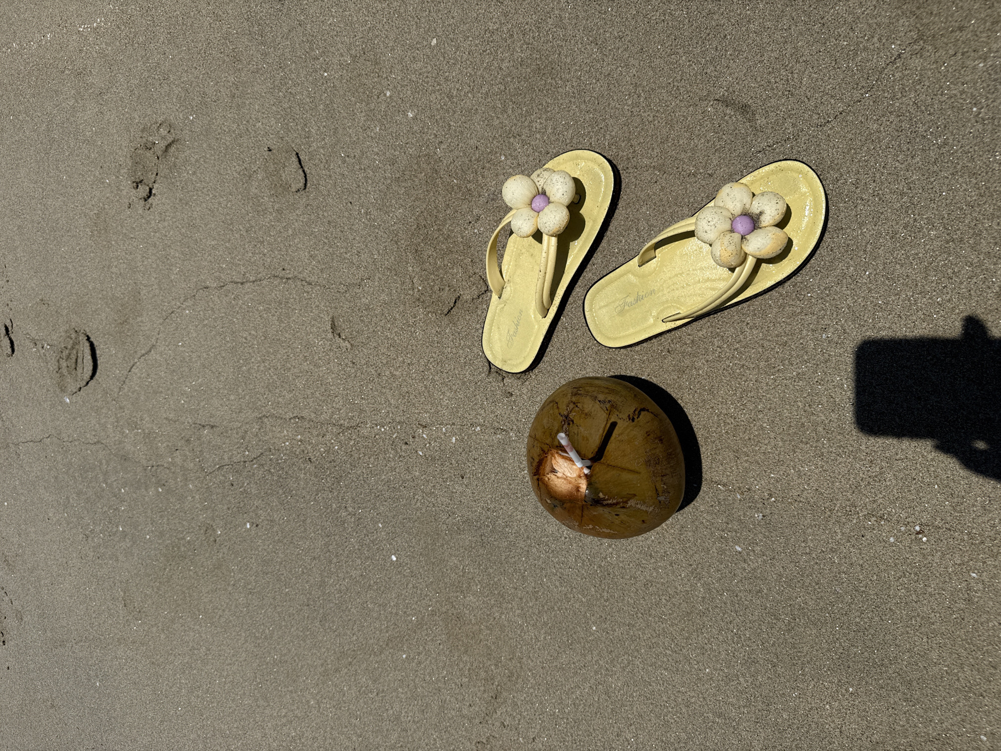 The image depicts a beach scene. A pair of yellow flip-flops with flower decorations and a drinking coconut are resting on the sand. Footprints are visible in the sand on the left side. A shadow is cast on the sand to the right. The location is presumed to be Đà Nẵng, Vietnam.