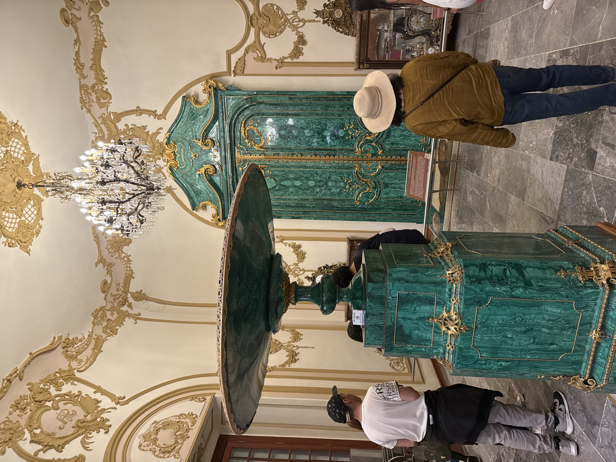 The image depicts an ornate interior space, likely a historical building or museum in Mexico City, Mexico. The focal point is a large, green malachite decorative water basin or fountain with intricate gold accents. Two people are in the scene: one wearing a brown jacket, blue jeans, and a hat, and another wearing a white shirt, gray pants, and a black cap. The first person appears to be looking at a large, green malachite door or wall feature that has gold accents. The other person is near the water basin.
The room features elaborate ceiling decorations in gold and white, including ornate moldings and a large chandelier. Other decorative elements include a tall clock or display cabinet. The floor is tiled in gray.