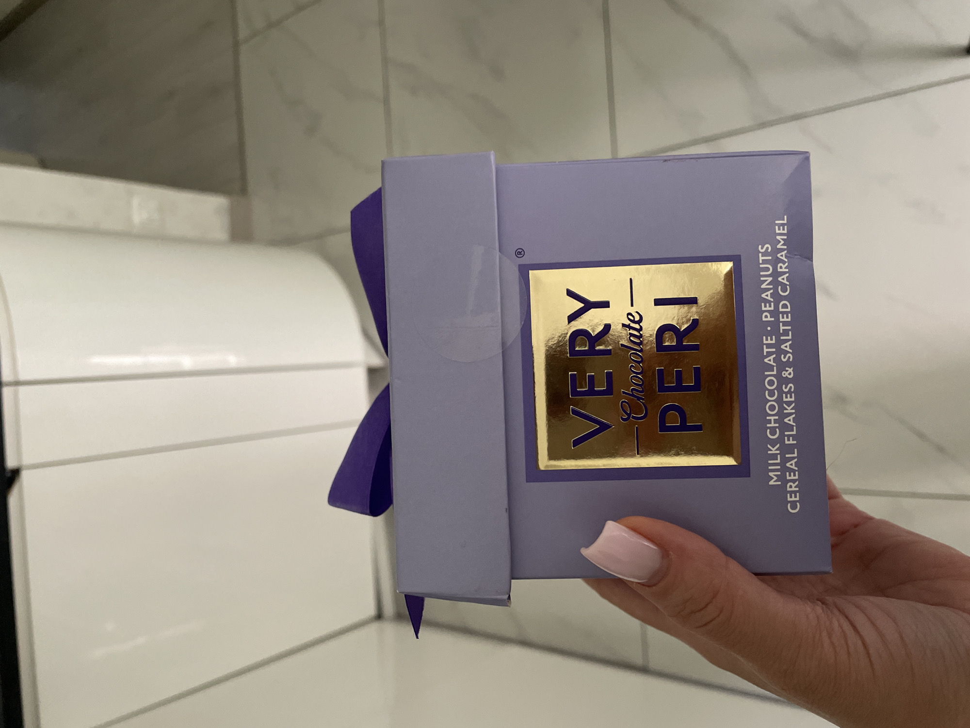 A hand is holding a lavender box of Very Chocolate Peri brand chocolates. The box is tied with a purple ribbon. The package states the chocolate includes milk chocolate, peanuts, cereal flakes, and salted caramel. The background appears to be a wall of white tiles and a curved, white object. The location is unknown and cannot be determined based on the provided image.